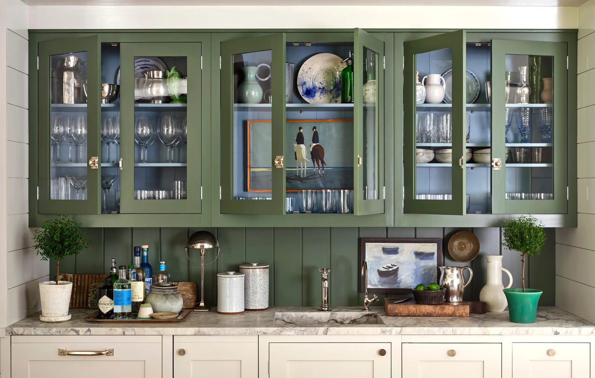 Sage green glass-front upper cabinets with light blue interiors sit above a white marble countertop with an undermount sink and green vertical backsplash.