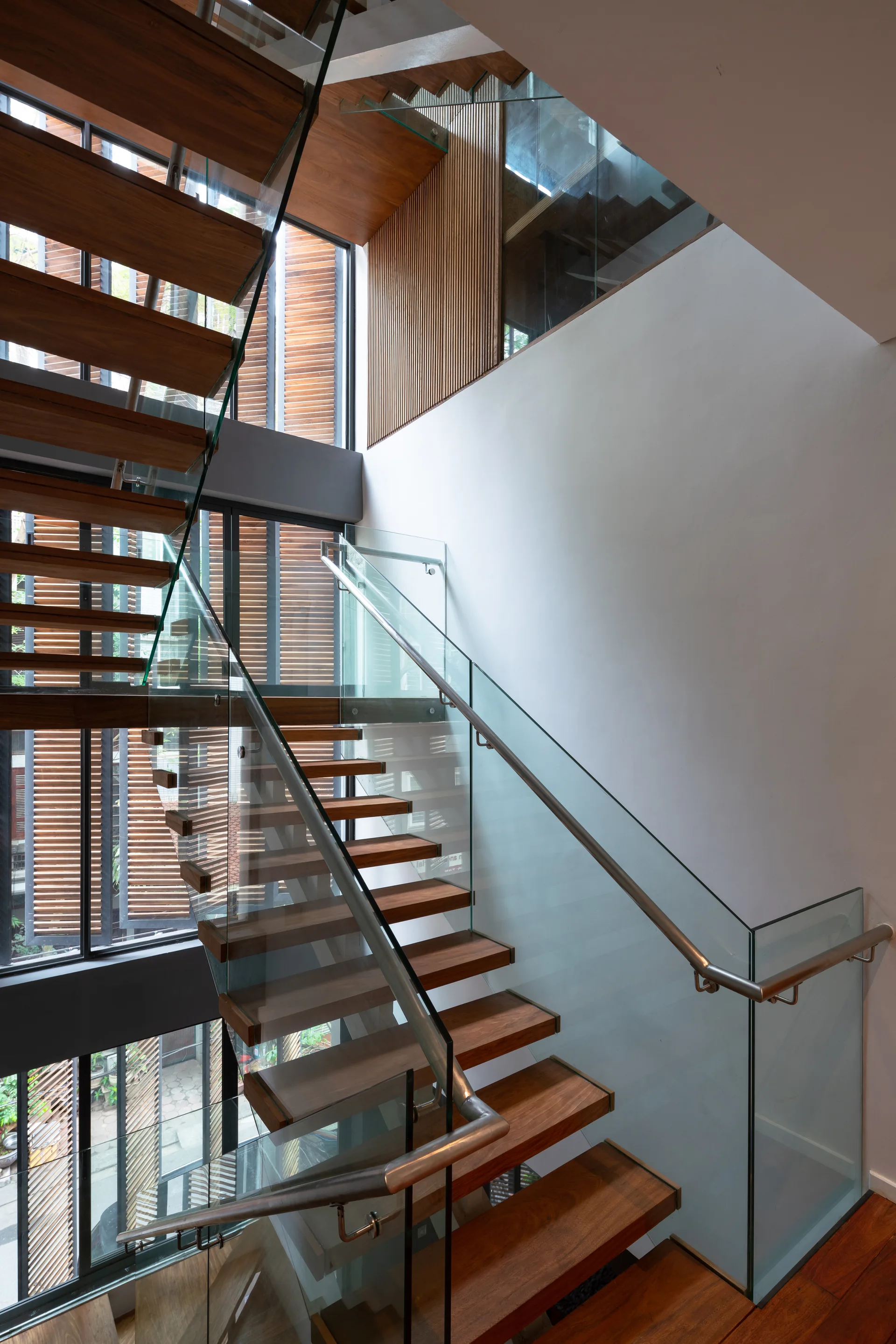 A contemporary floating staircase with natural wood treads and clear glass railings framed by white walls and a large window.