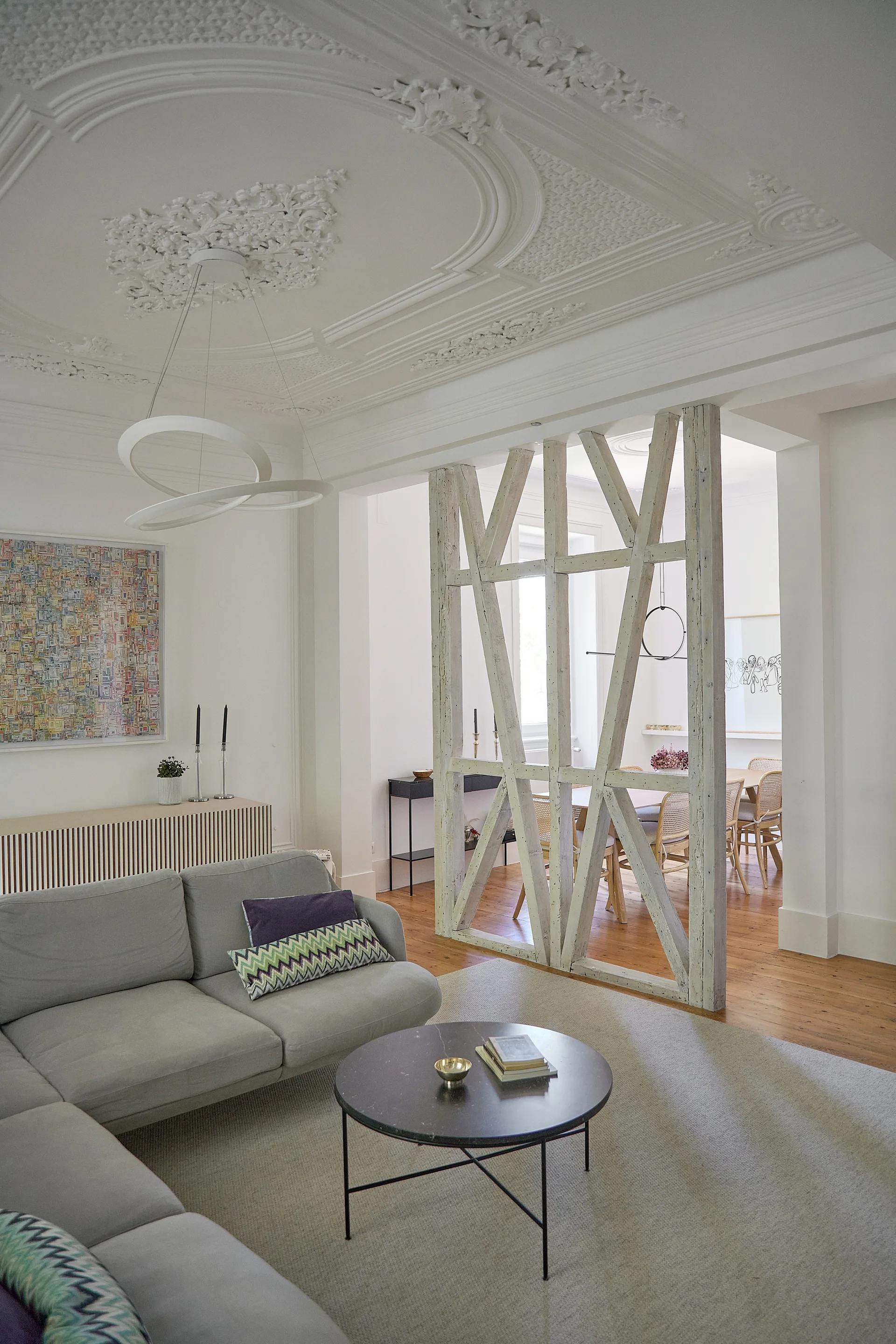 A contemporary living room features a grey sectional, a dark round coffee table, a rustic wood divider, and intricate white ceiling plaster.