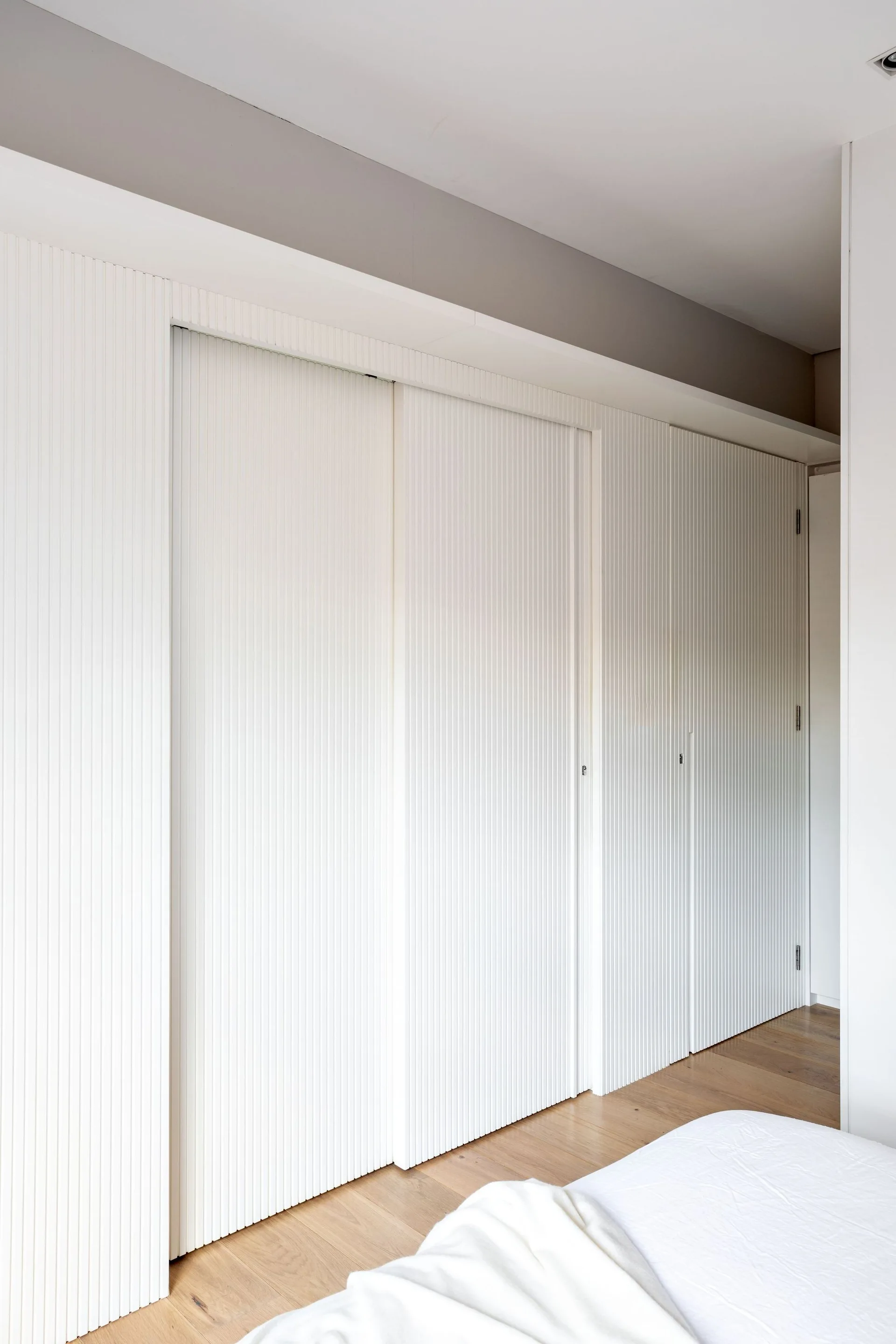 Floor-to-ceiling white sliding closet doors with vertical slats next to a light wood floor and a corner of a bed in a minimalist bedroom.