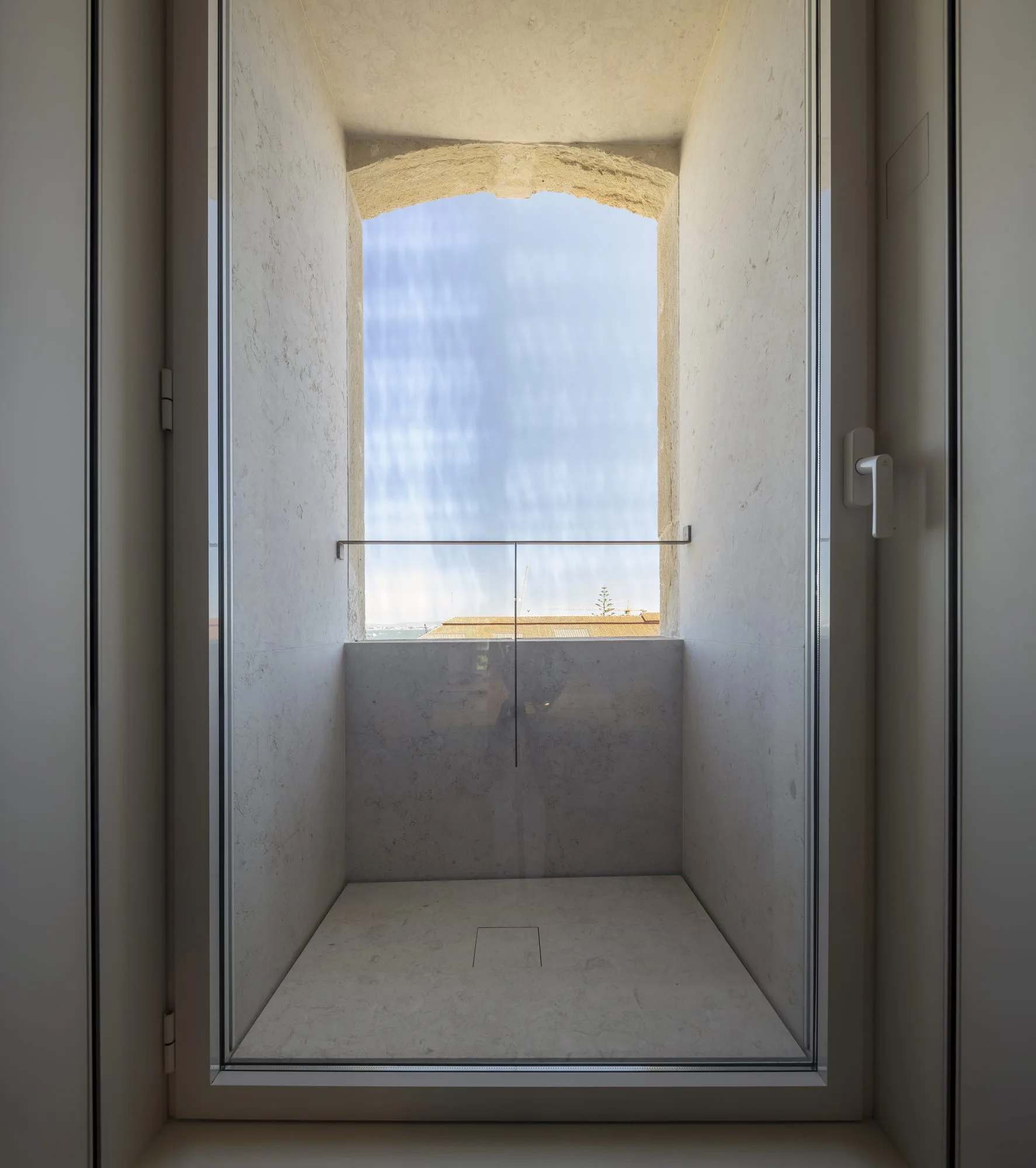 A deep off-white stone window niche features an arched stone header and an integrated glass balustrade looking out toward a blue sky.