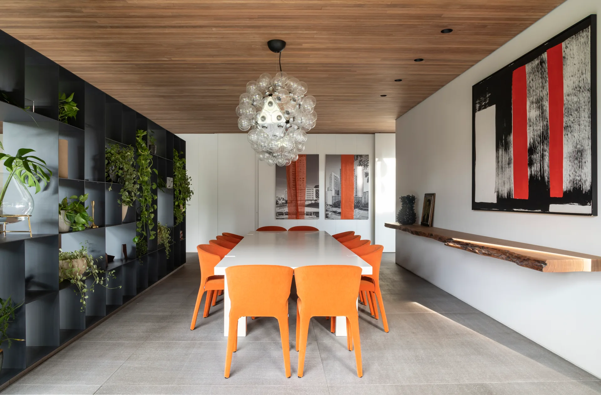 A modern dining room features a white table surrounded by orange chairs under a spherical bubble chandelier and a warm wood-plank ceiling.