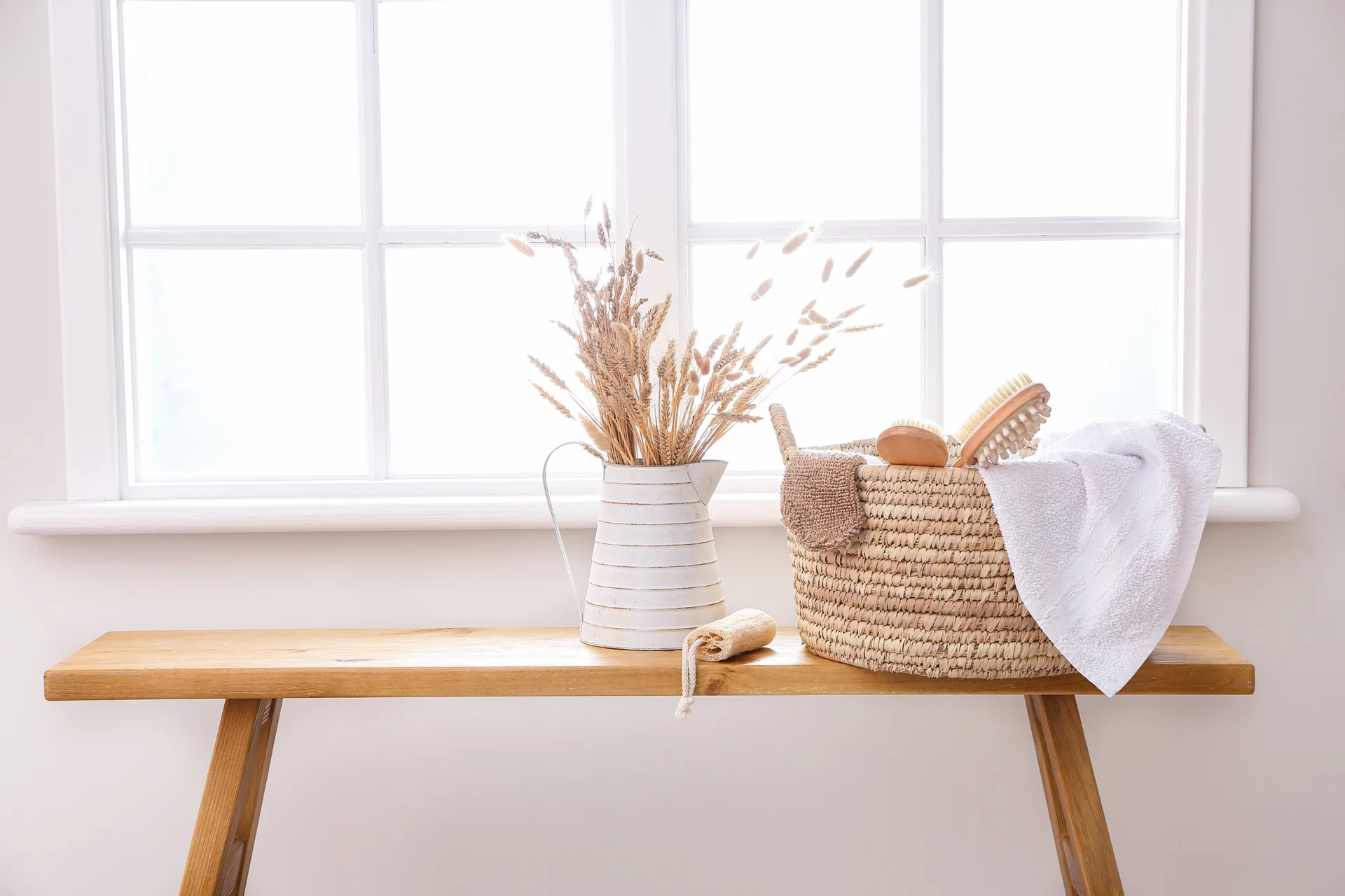 A natural wood bench sitting below a white window holds a woven basket of spa brushes and towels next to a white pitcher of dried wheat.