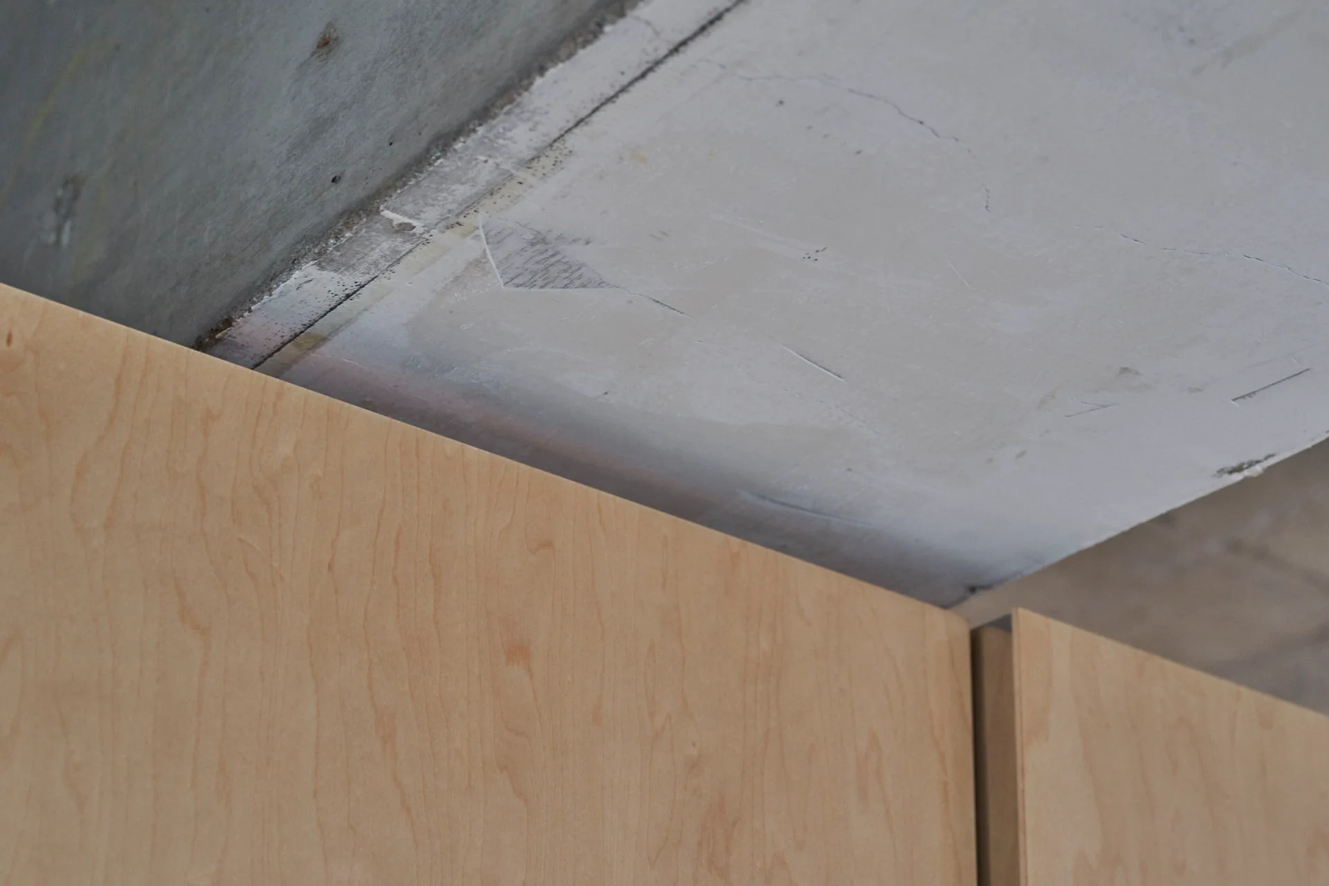 A close-up of the upper edge of light wood cabinetry meeting an unfinished, textured grey concrete ceiling.