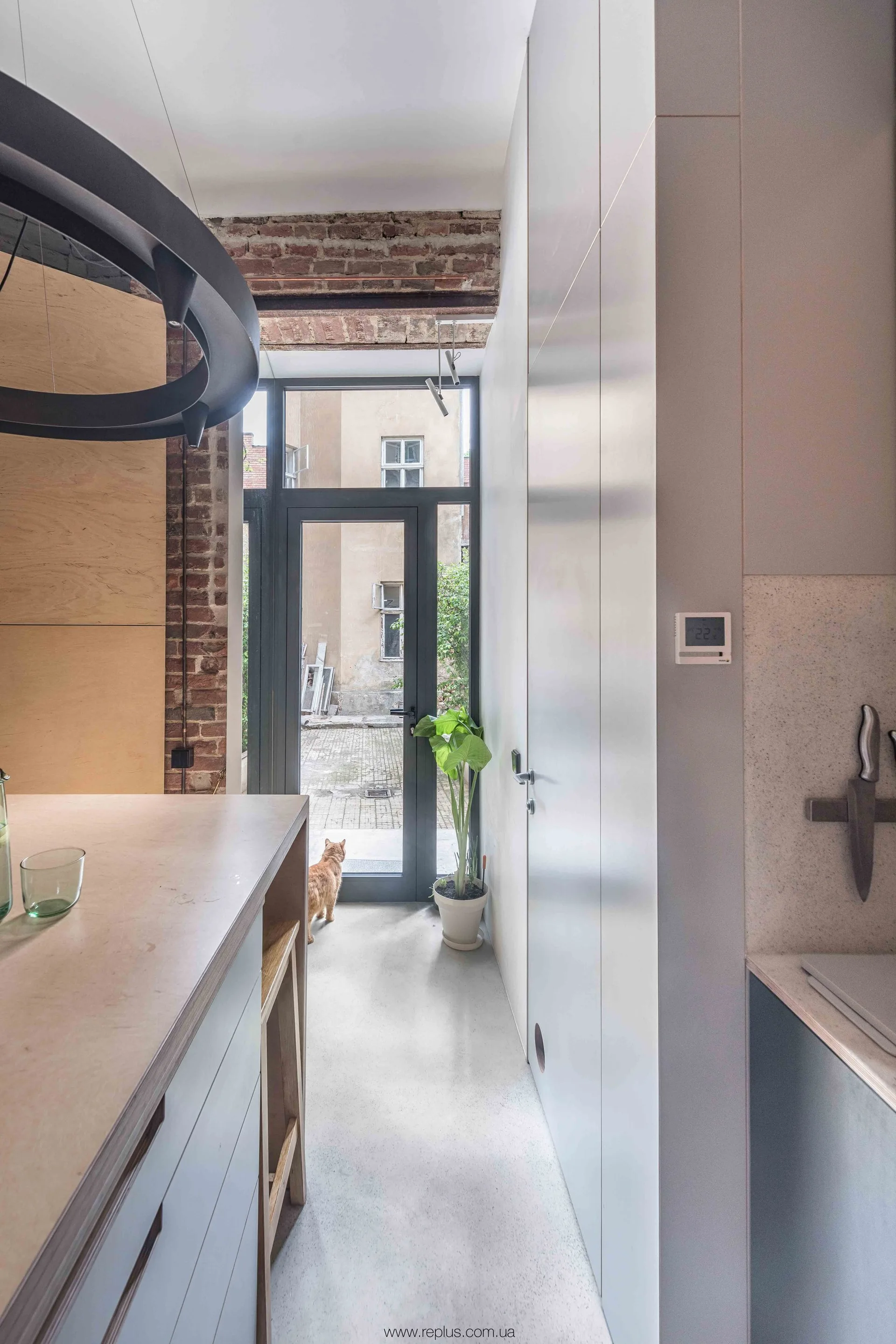 A narrow grey kitchen features floor-to-ceiling handleless cabinets, a light wood island, and a glass back door under exposed brick.