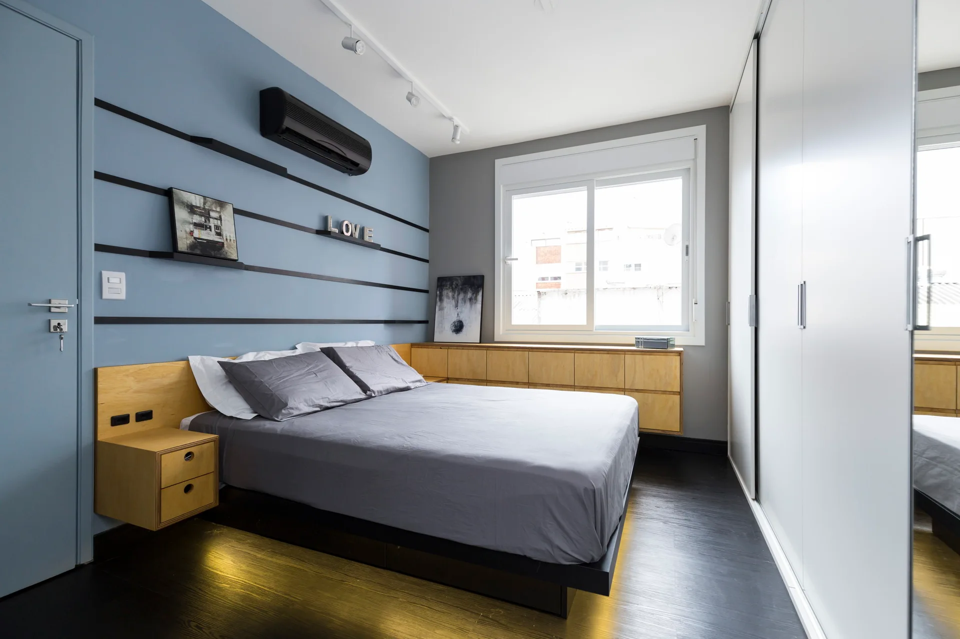 A modern bedroom features a platform bed with grey sheets, a blue accent wall with thin black stripes, and low light-wood cabinets under the window.
