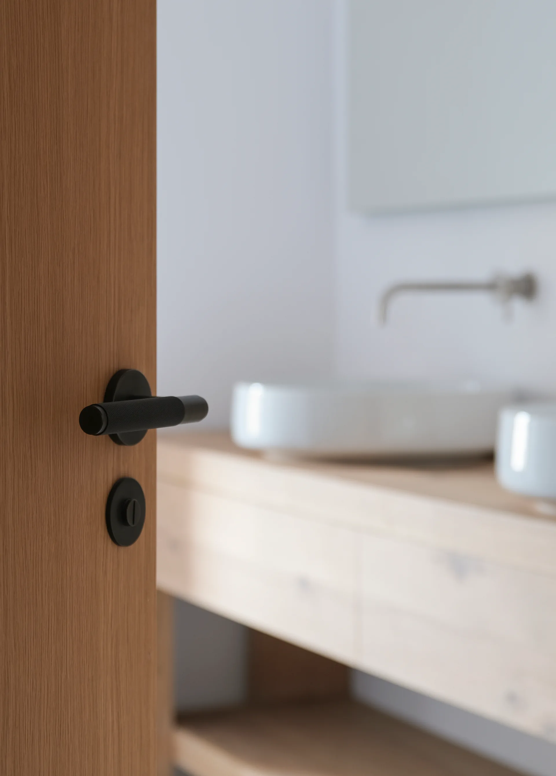 A close-up of a wood-grained door with a black knurled handle, with a blurry bathroom vanity and sink in the background.