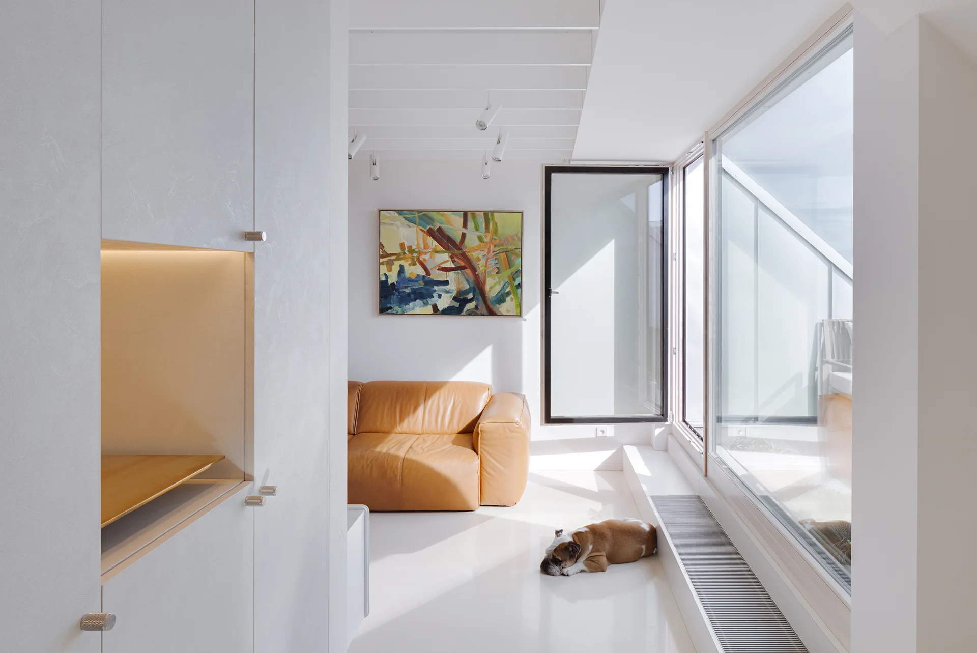 A minimalist white living room features a tan leather sofa, large windows, white cabinetry with a lit niche, and a dog lying on the floor.