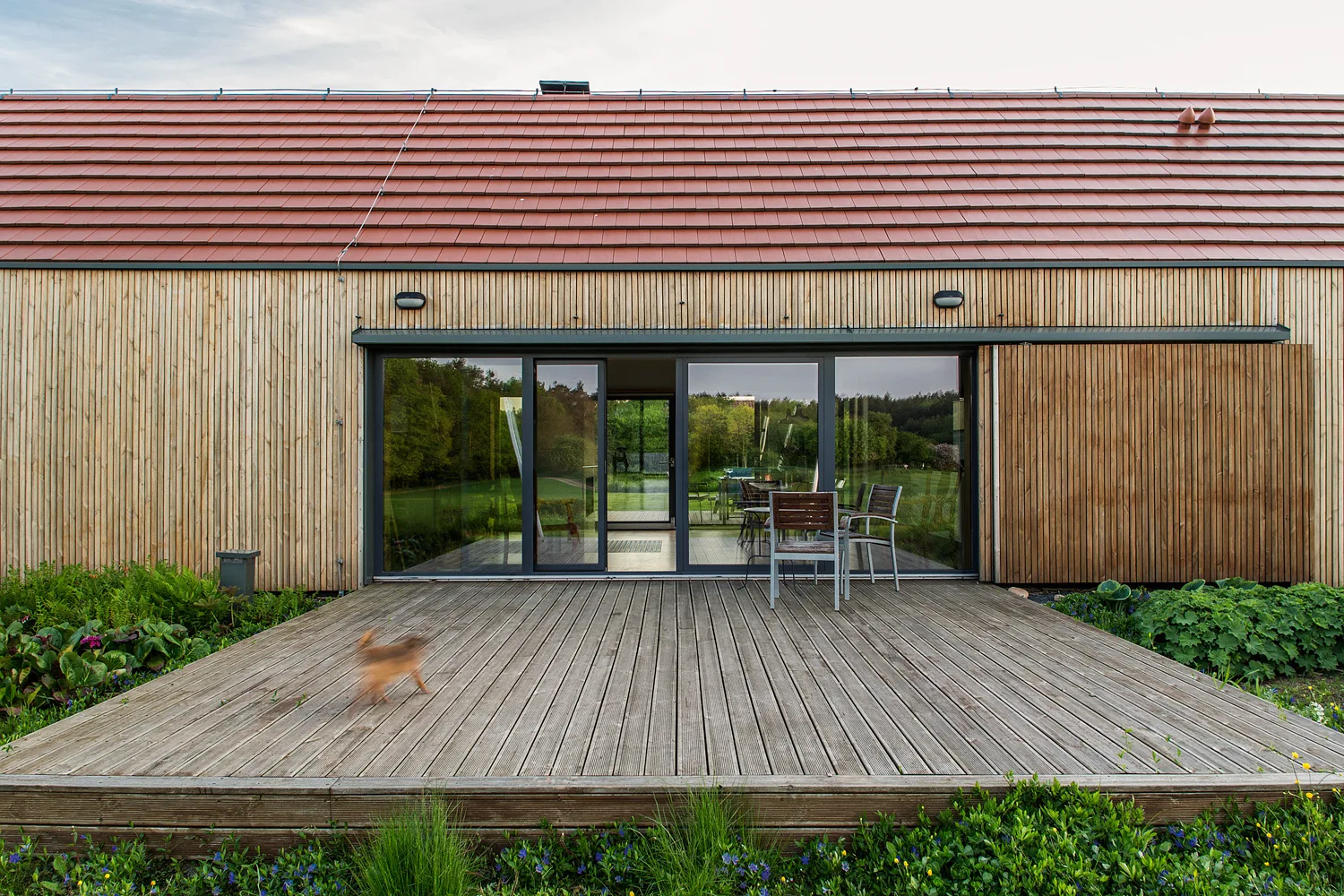 A modern house exterior featuring vertical wood slatted siding, a red tile roof, and a large glass sliding door opening onto a weathered wooden deck.