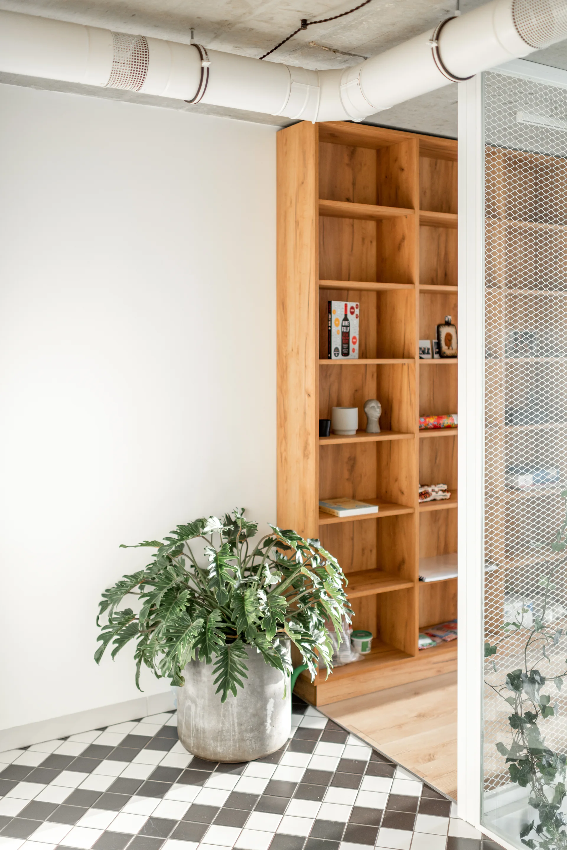 A modern transitional space features checkered black and white floor tiles, a tall wooden bookshelf, a large potted plant, and a white wire mesh partition.