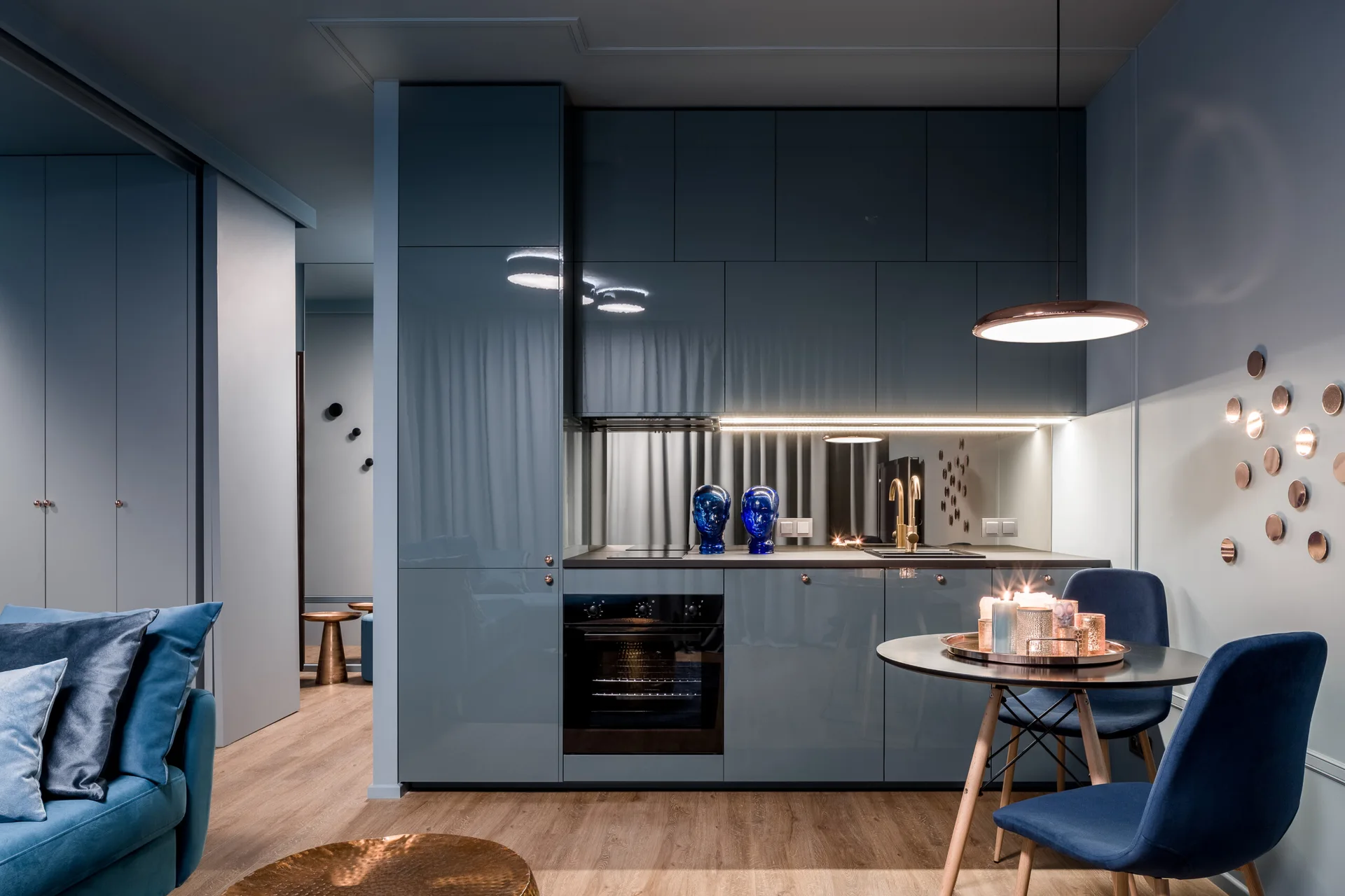 A contemporary kitchen features high-gloss blue-grey cabinets, a mirrored backsplash with under-cabinet lighting, and a small round dining table.