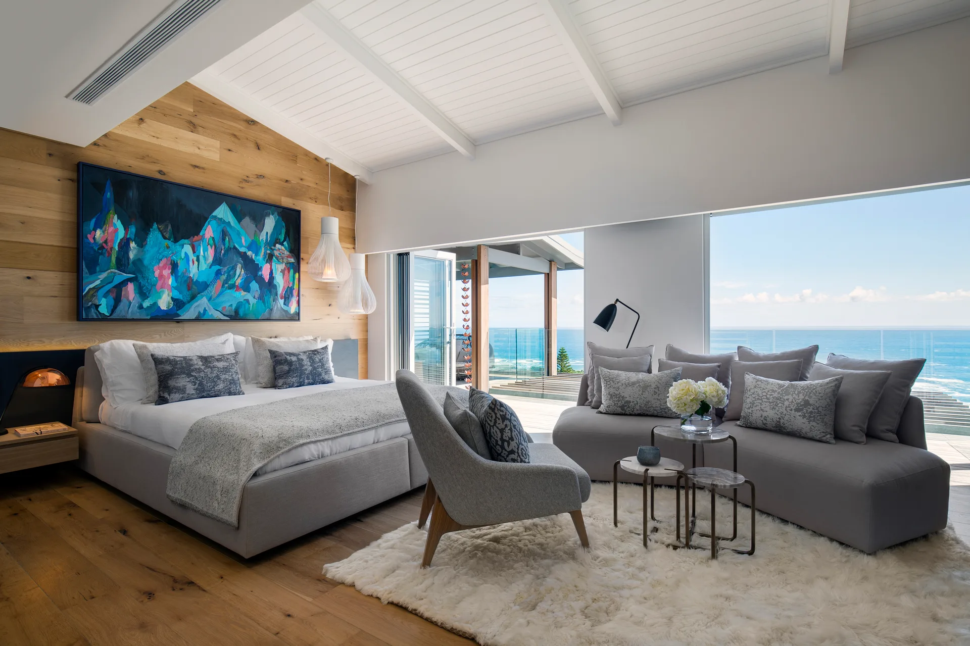 A spacious bedroom with a wood-plank accent wall, a large bed, and a grey sectional sofa facing floor-to-ceiling windows with an ocean view.