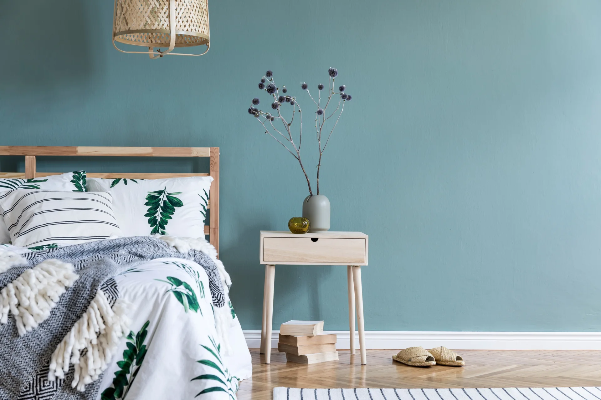 A bedroom features a light wood bed with botanical leaf-print bedding and a matching nightstand against a solid teal accent wall on herringbone floors.