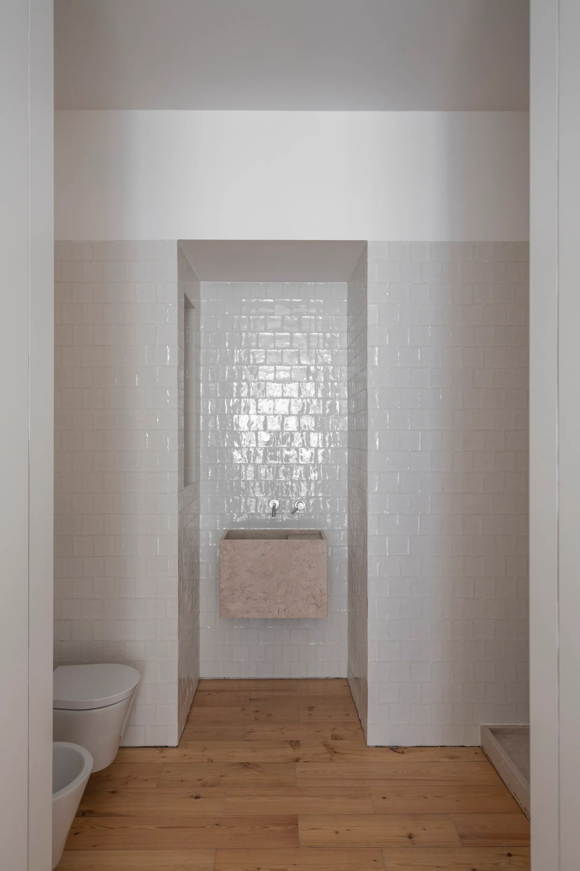 A minimalist bathroom with a floating stone basin set in a tiled alcove, featuring white square wall tiles and natural wood plank flooring.