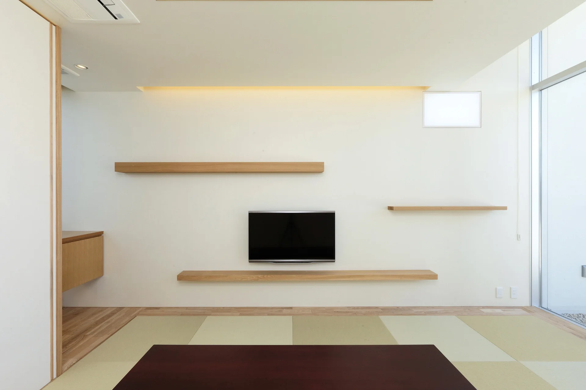 A wall-mounted television is framed by asymmetrical wooden floating shelves in a minimalist room with tatami mat flooring.