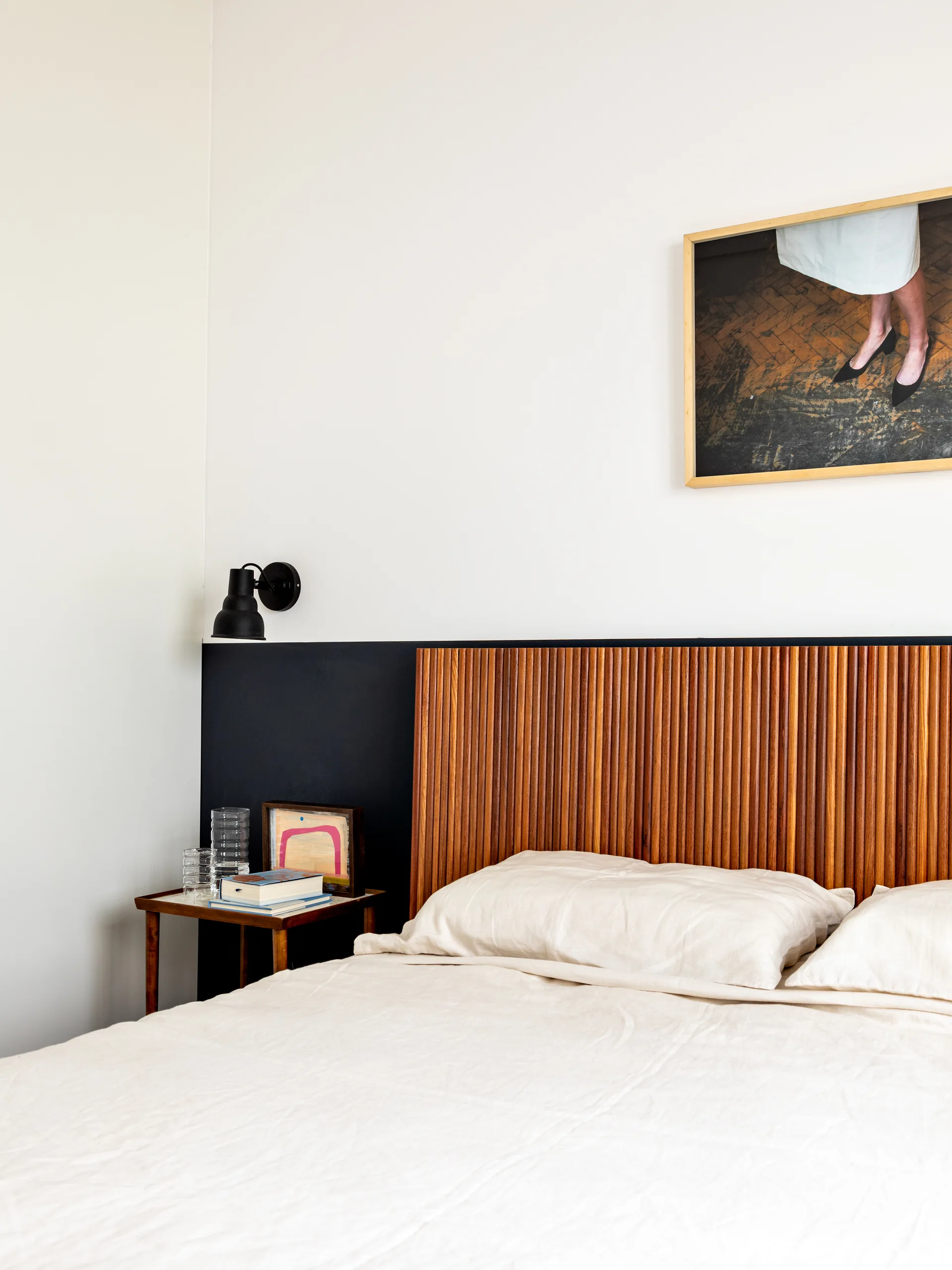 A minimalist bedroom featuring a fluted wood headboard against a two-tone black and white wall, next to a simple wood nightstand.