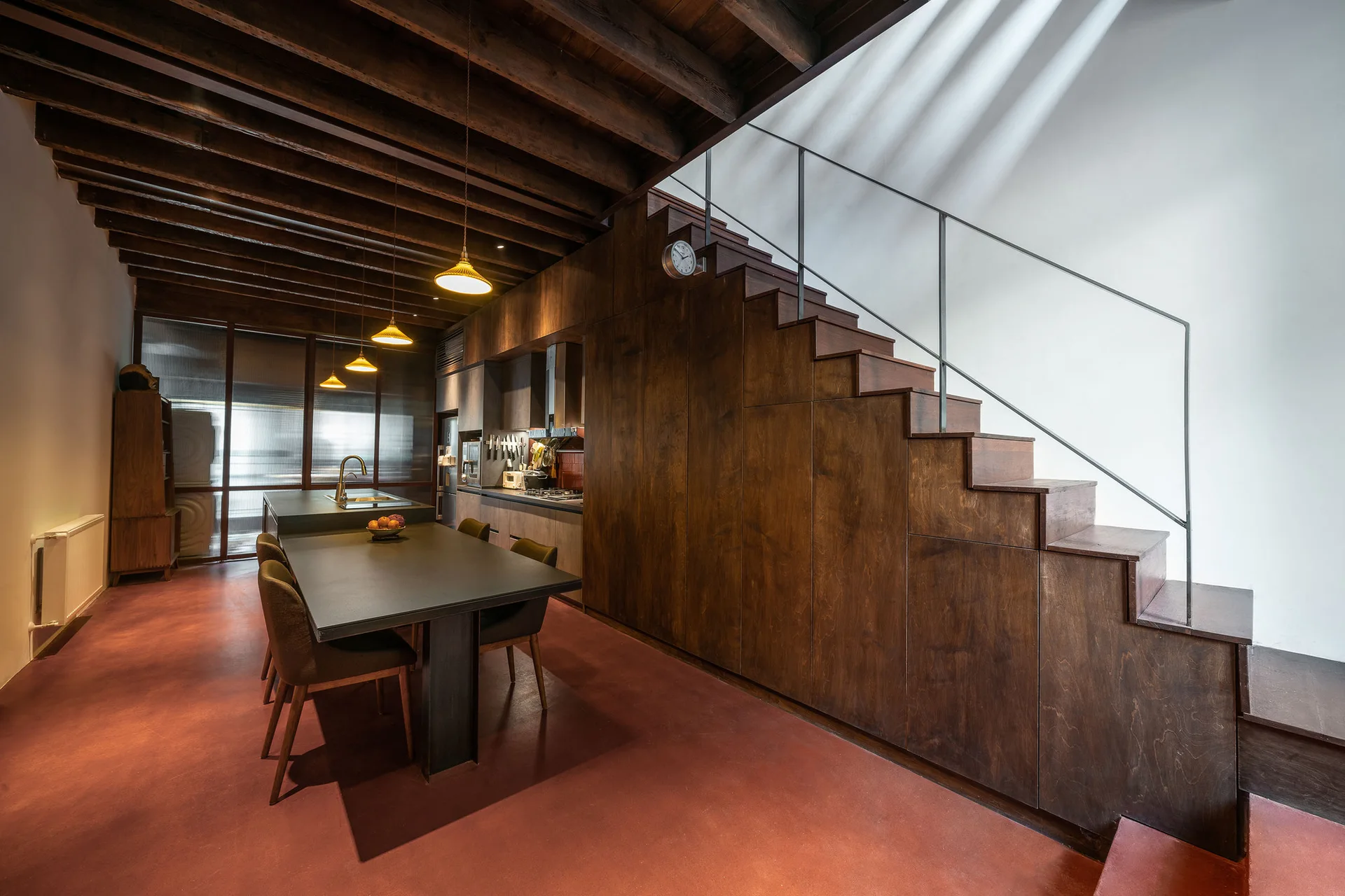 A modern kitchen featuring a dark wood integrated staircase with built-in storage, a matching dining island, and a distinctive red floor.