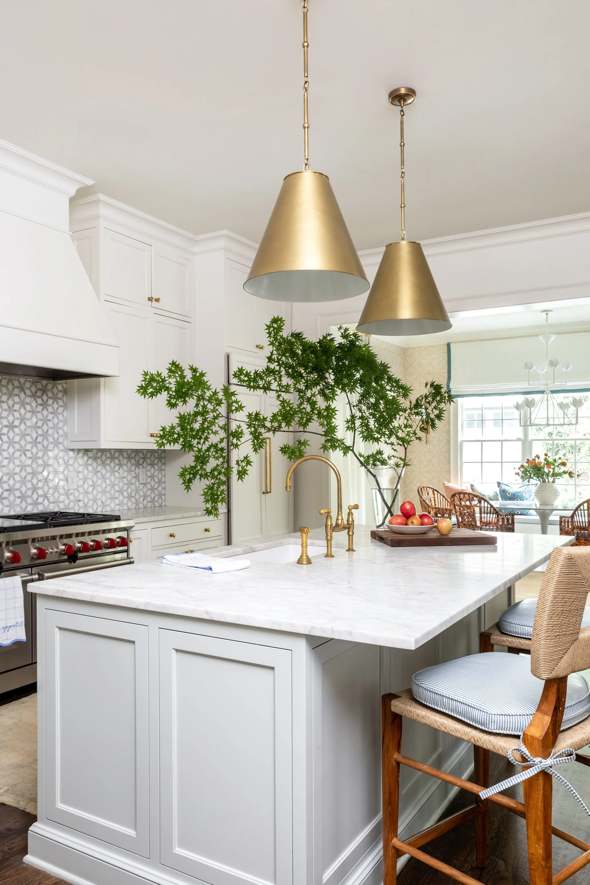 A bright kitchen features a white marble-topped island with a brass faucet, woven barstools, two brass pendant lights, and a geometric tile backsplash.