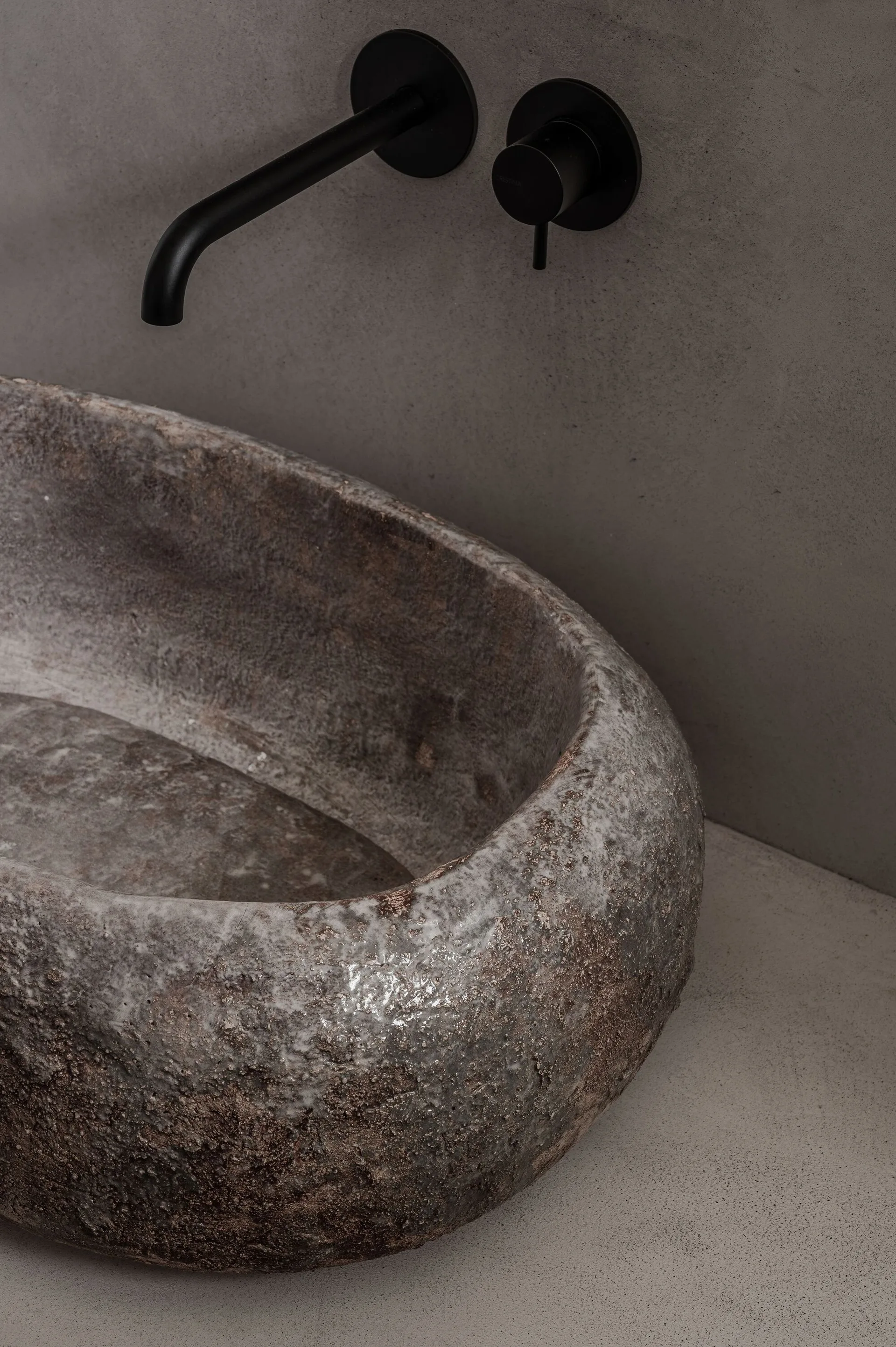 A textured brown stone vessel sink sits on a grey countertop beneath wall-mounted matte black plumbing fixtures on a smooth grey wall.