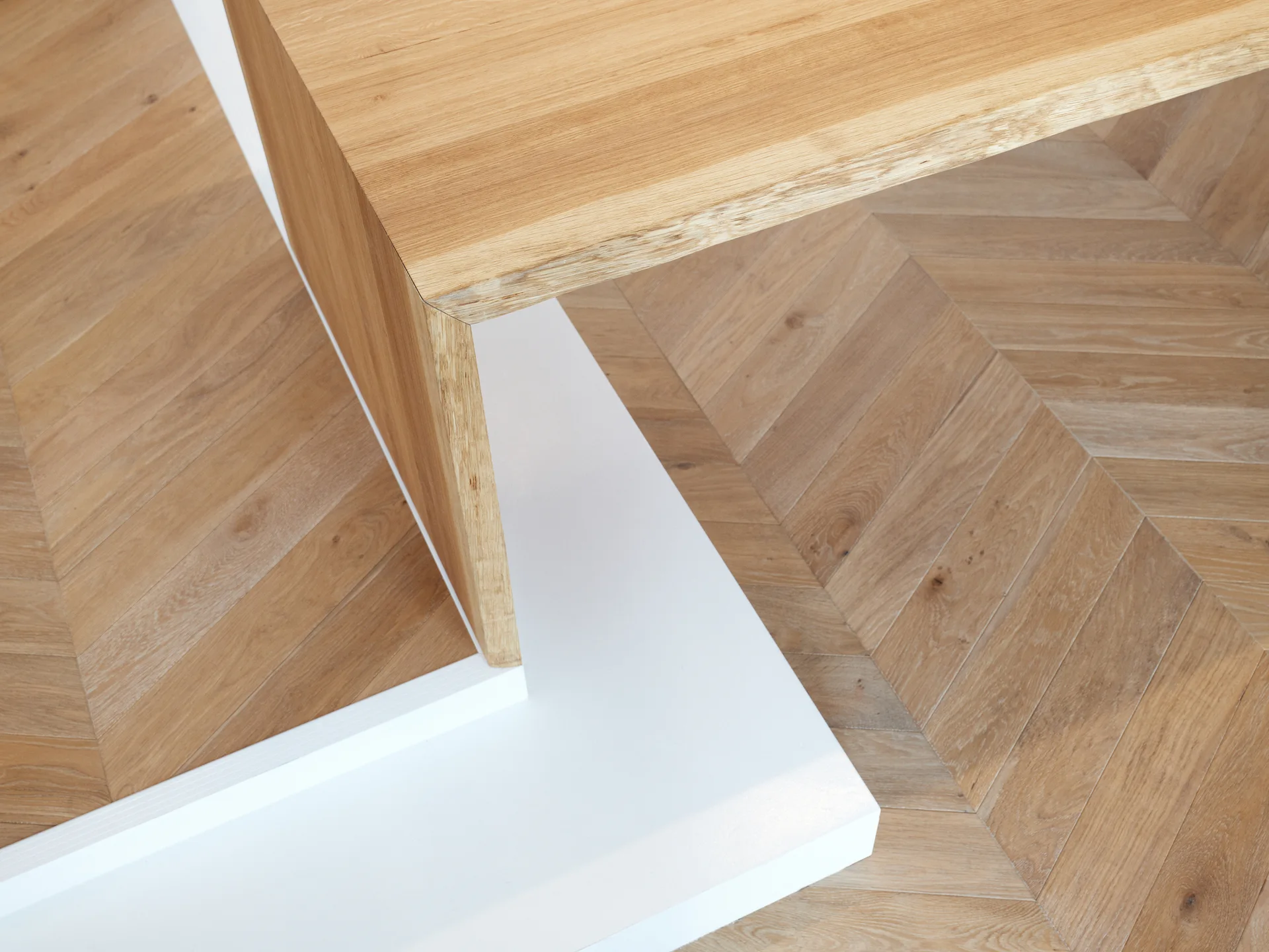 A close-up view of an angular light oak built-in table meeting a solid white base over a chevron wood plank floor.