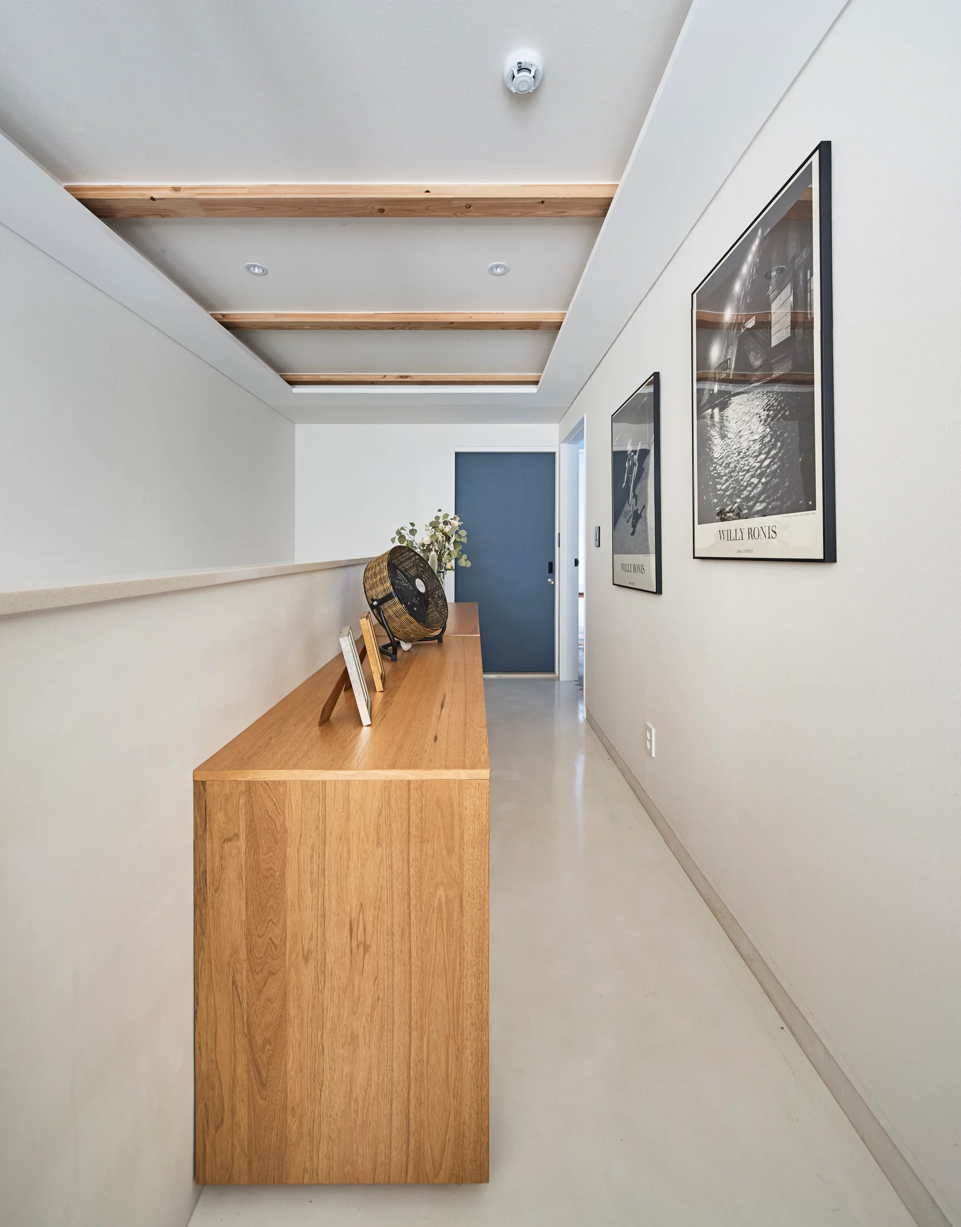 A minimalist hallway features a long wooden console table, exposed wood ceiling beams, and framed black and white art leading to a blue door.