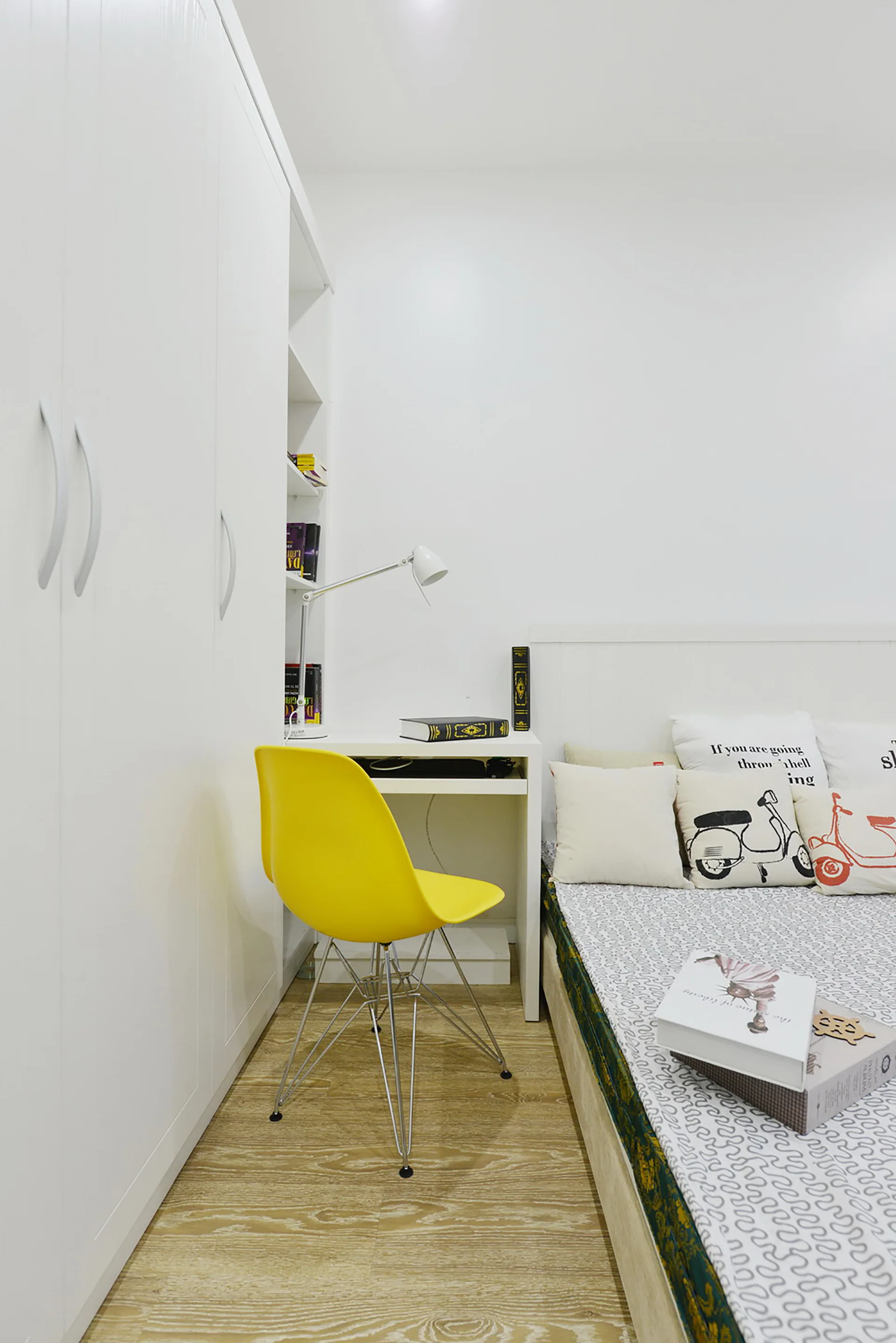 A modern bedroom with a white wardrobe, an integrated desk, a bright yellow molded plastic chair, and a bed with decorative graphic pillows.