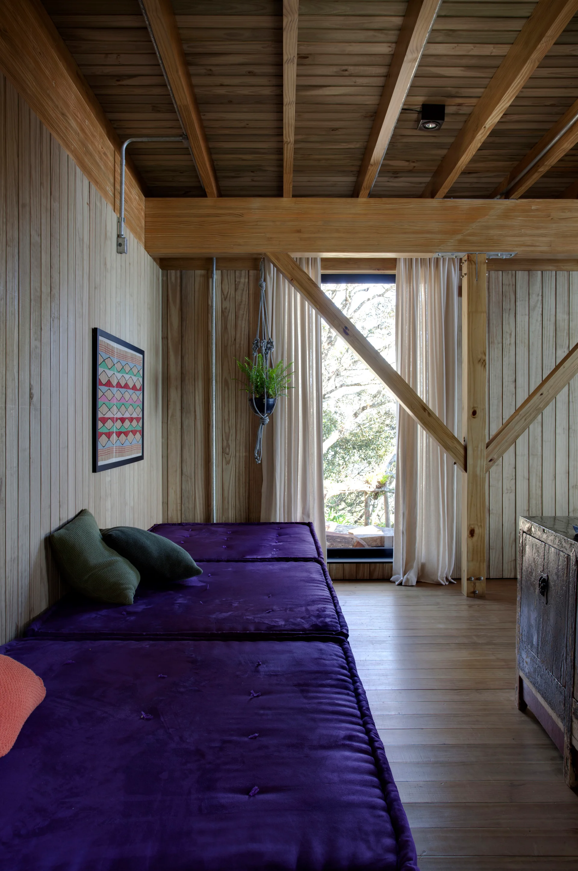 A light wood paneled room features a long purple velvet floor mattress with throw pillows under an exposed beam ceiling.