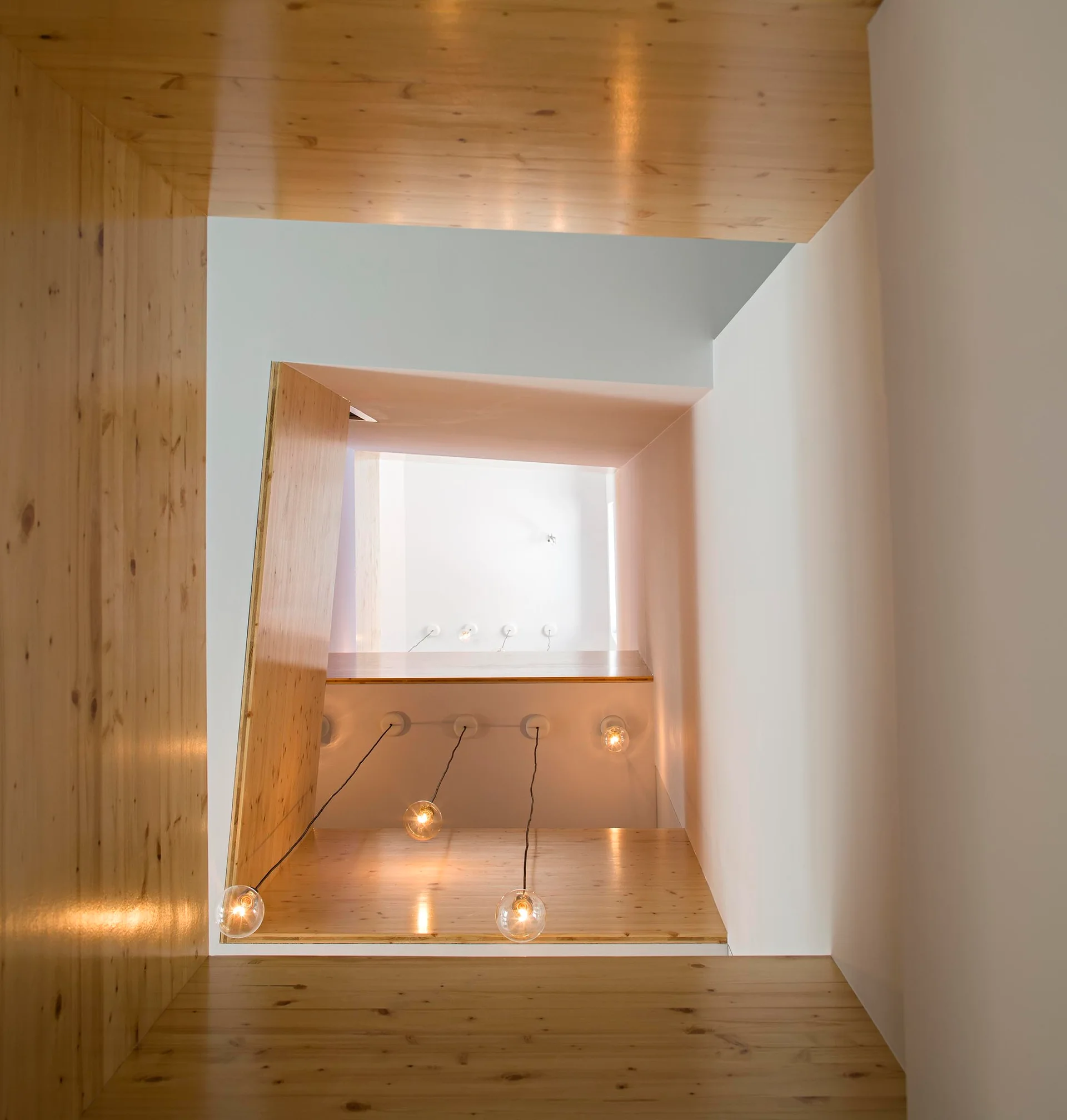 A modern minimalist stairwell void with light knotty pine paneling, white walls, and clear glass pendant lights hanging at staggered heights.