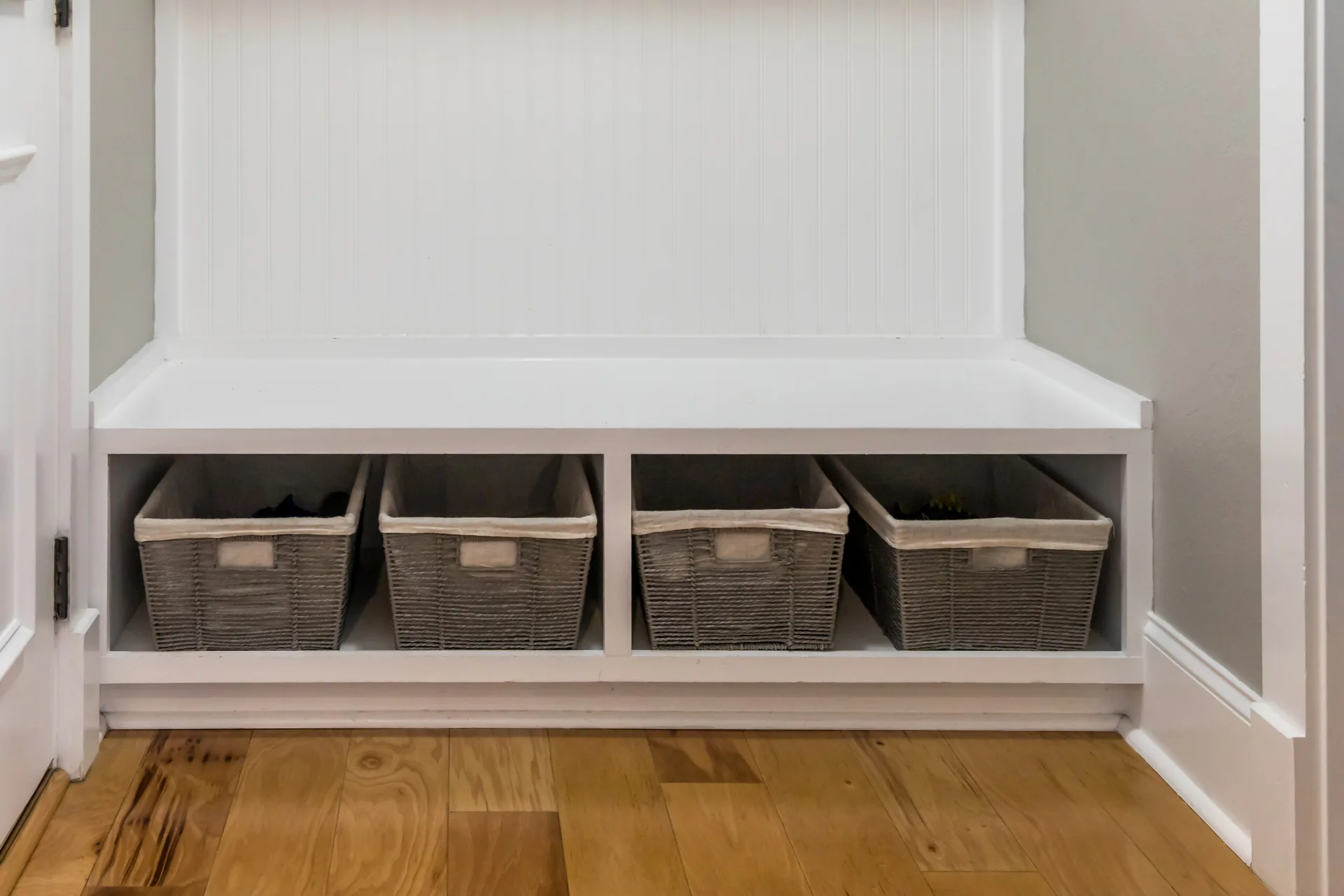 A white built-in entryway bench features four grey woven storage baskets beneath the seat, set against a white beadboard wall.