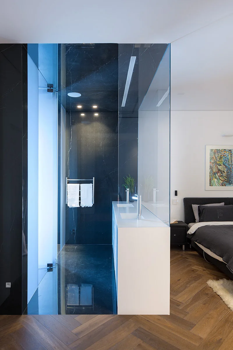 A bedroom featuring herringbone wood flooring next to a glass-enclosed bathroom with black marble walls and a white double vanity.