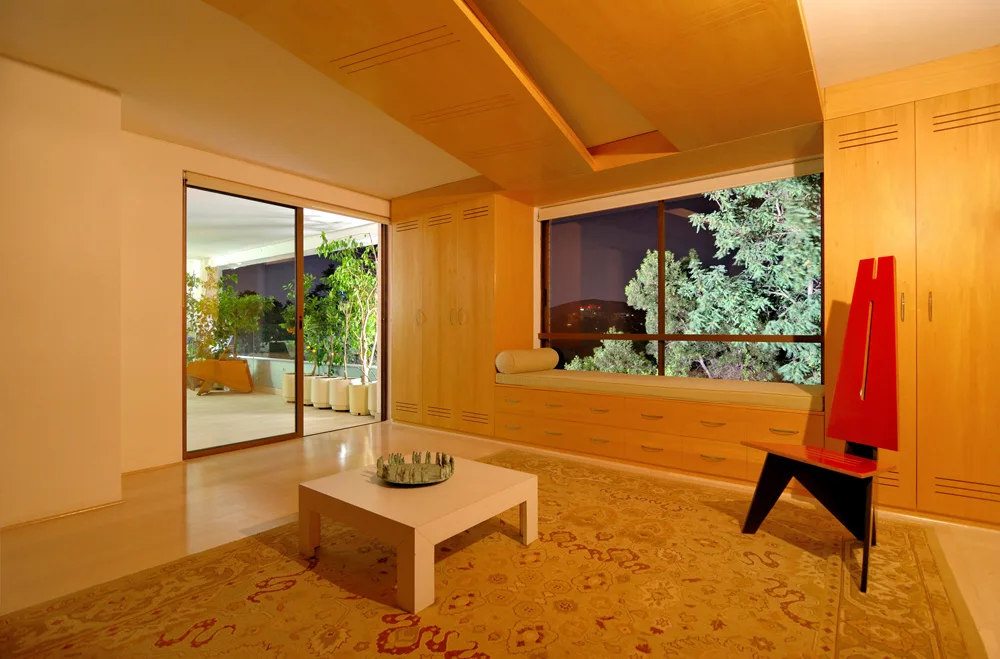A contemporary room features light wood built-in cabinets framing a window seat, a matching wood ceiling panel, a patterned rug, and a red accent chair.