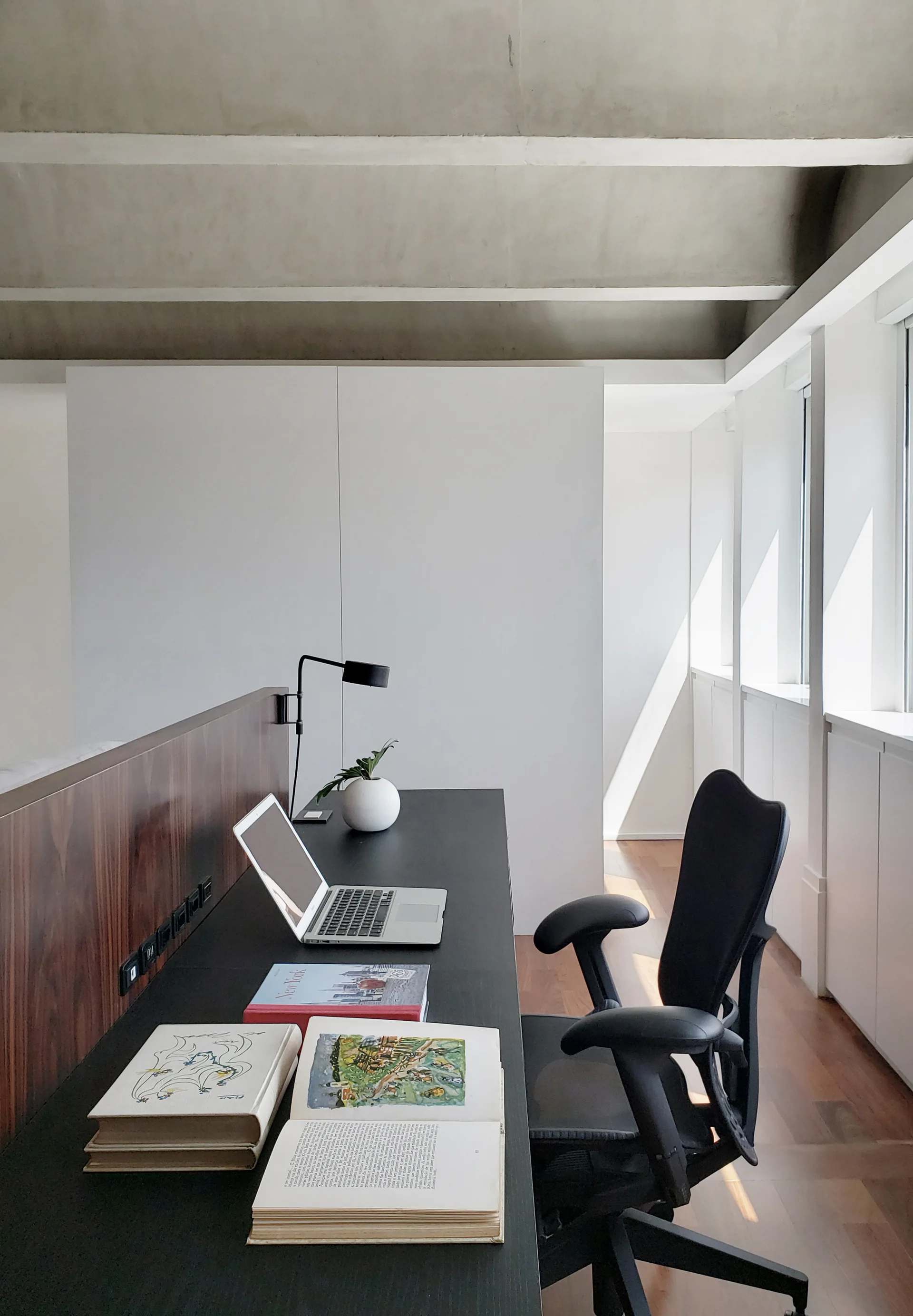 A minimalist home office features a dark wood-backed desk with a laptop, a black ergonomic chair, and a high grey concrete ceiling with exposed beams.