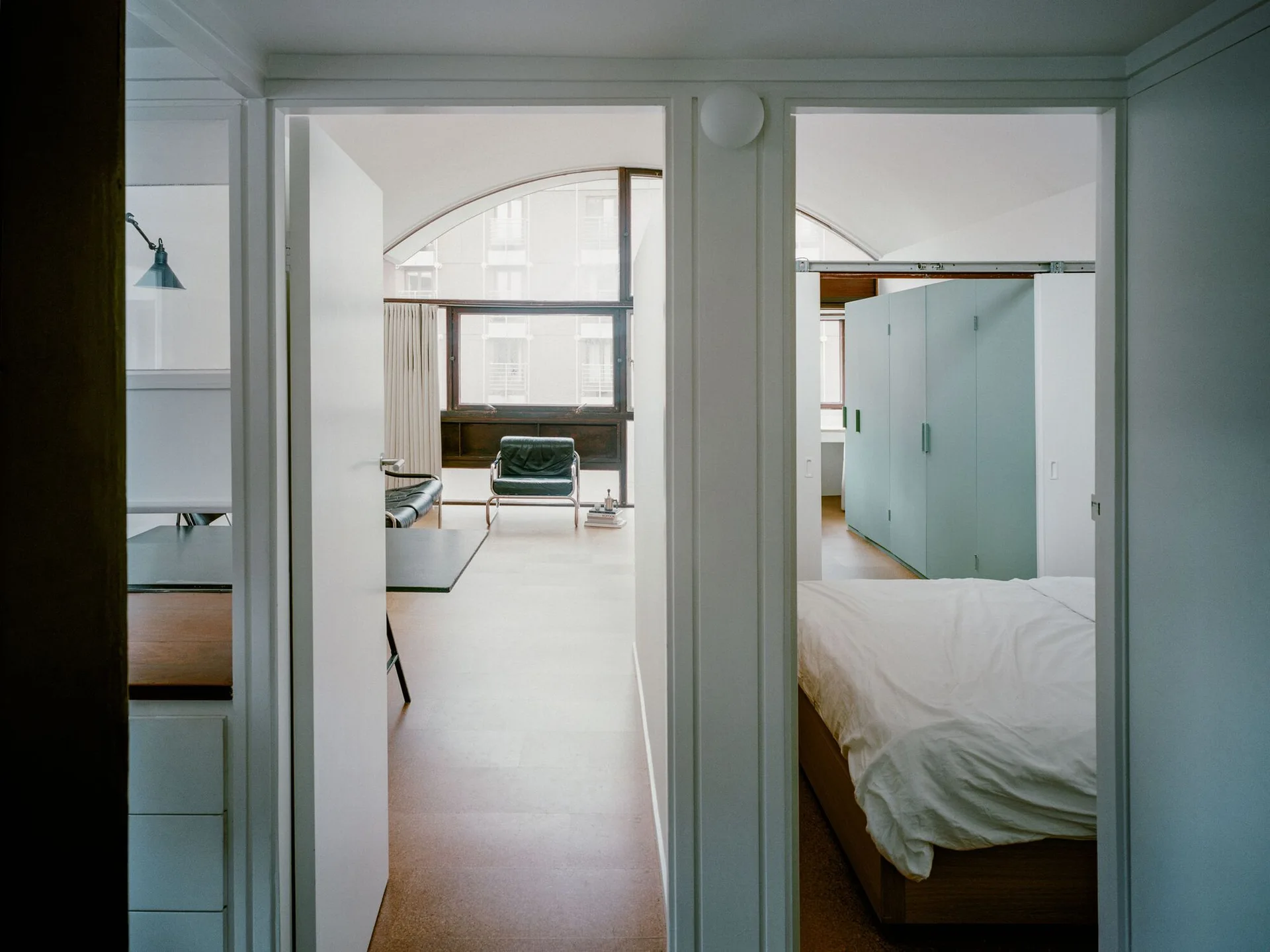 A modern bedroom view showing a white bed and a sage green wardrobe, with an adjacent living area and arched window visible in the background.