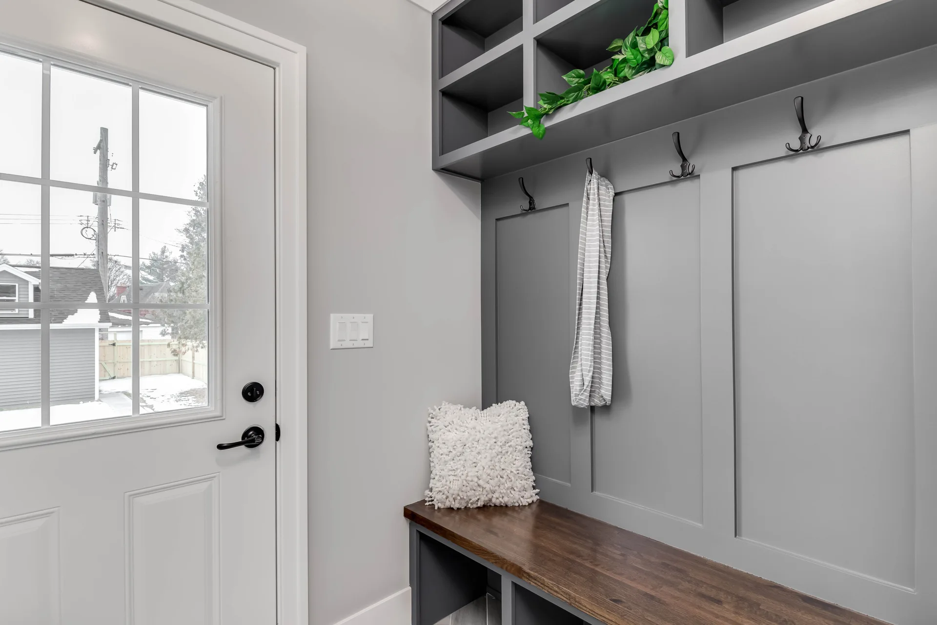 A mudroom entryway featuring a grey built-in storage unit with a wooden bench, black coat hooks, upper cubbies, and a white glass-paneled door.
