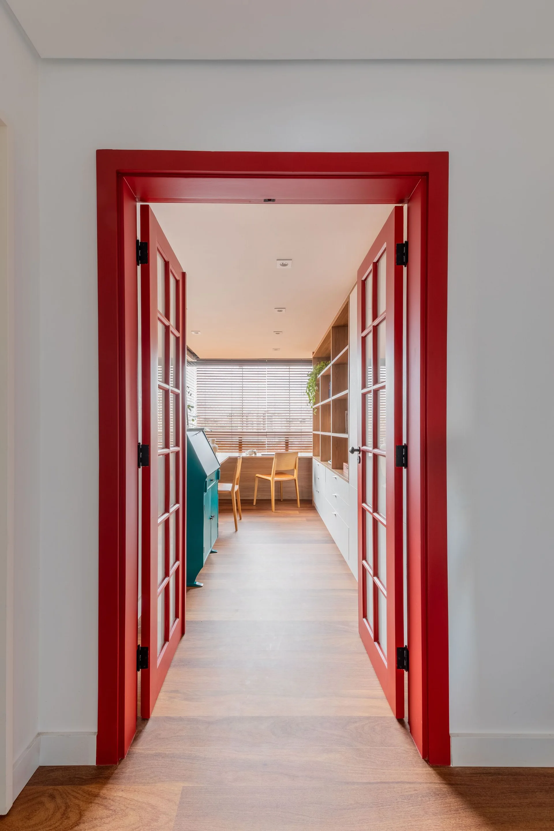 A view through bright red painted glass double doors into a home office with wood floors, built-in shelving, and a desk.