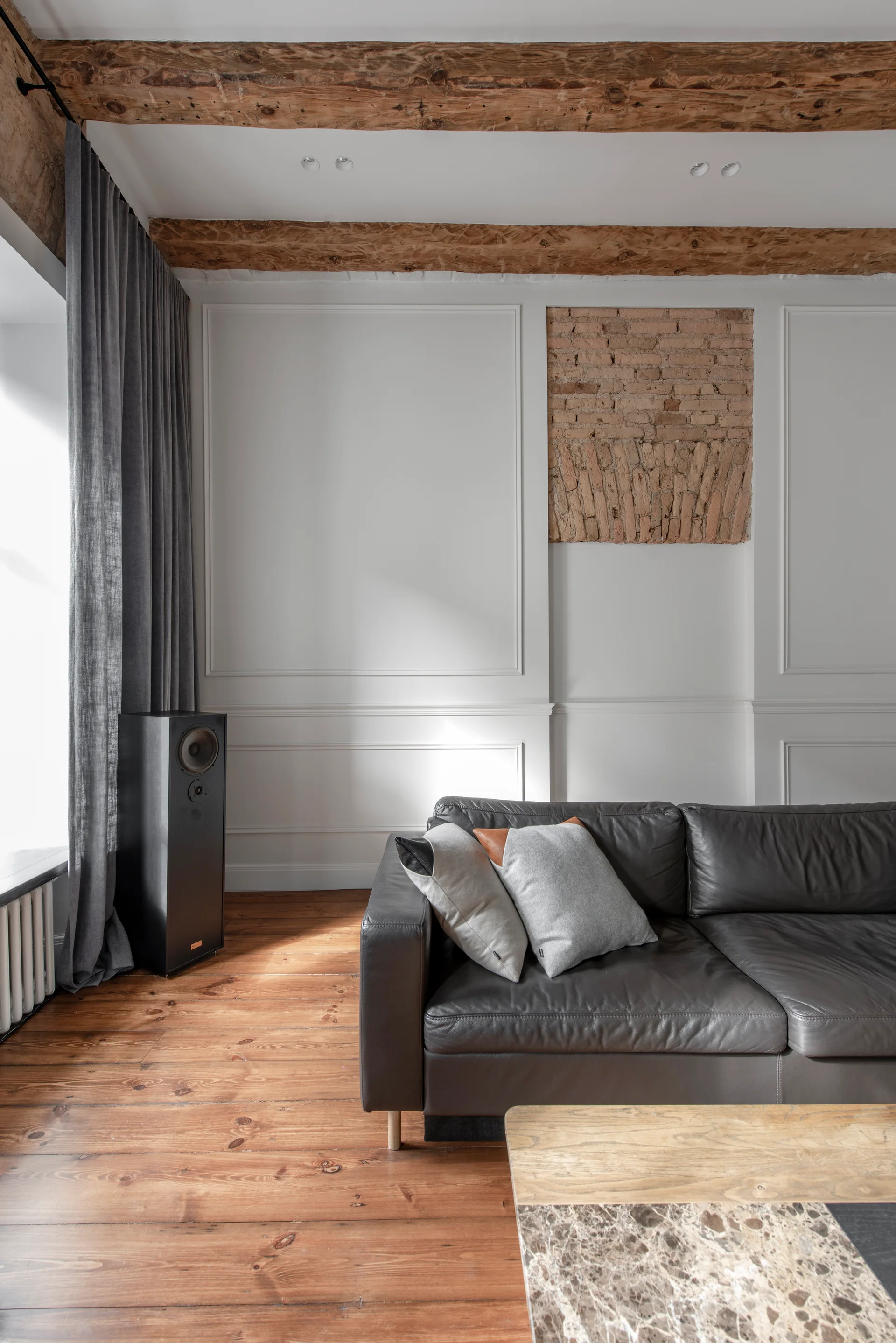 A living room with a dark leather sofa, exposed wooden ceiling beams, white paneled walls, and a small rectangular section of exposed brick.
