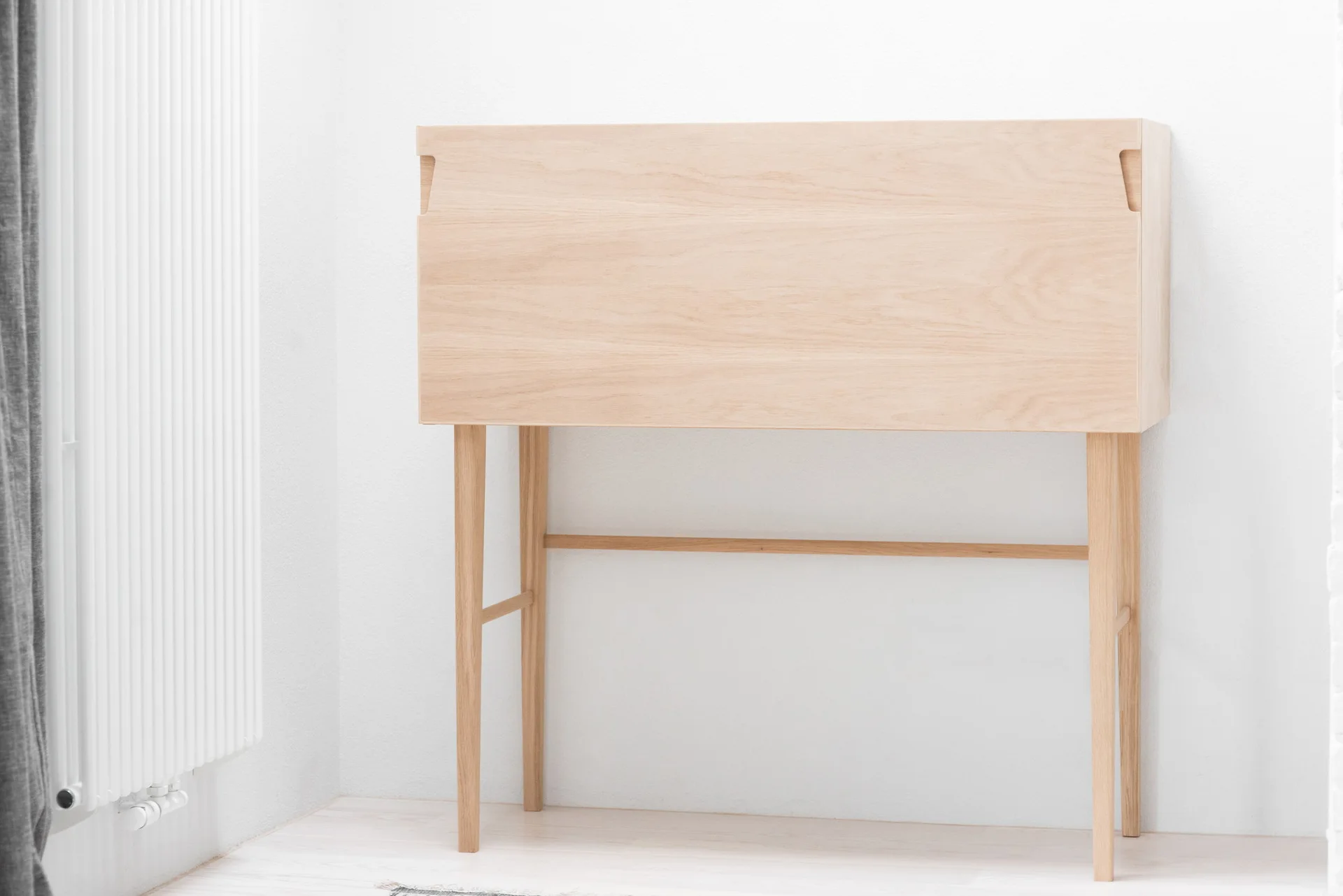 A minimalist light wood secretary desk with tapered legs stands against a white wall next to a vertical radiator.