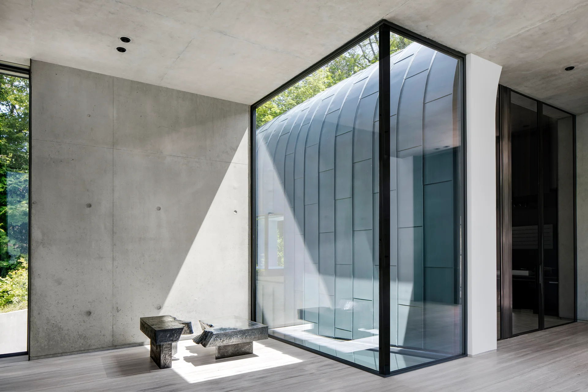 A minimalist room features raw concrete walls, light wood floors, a dark sculptural side table, and a large floor-to-ceiling glass corner window.