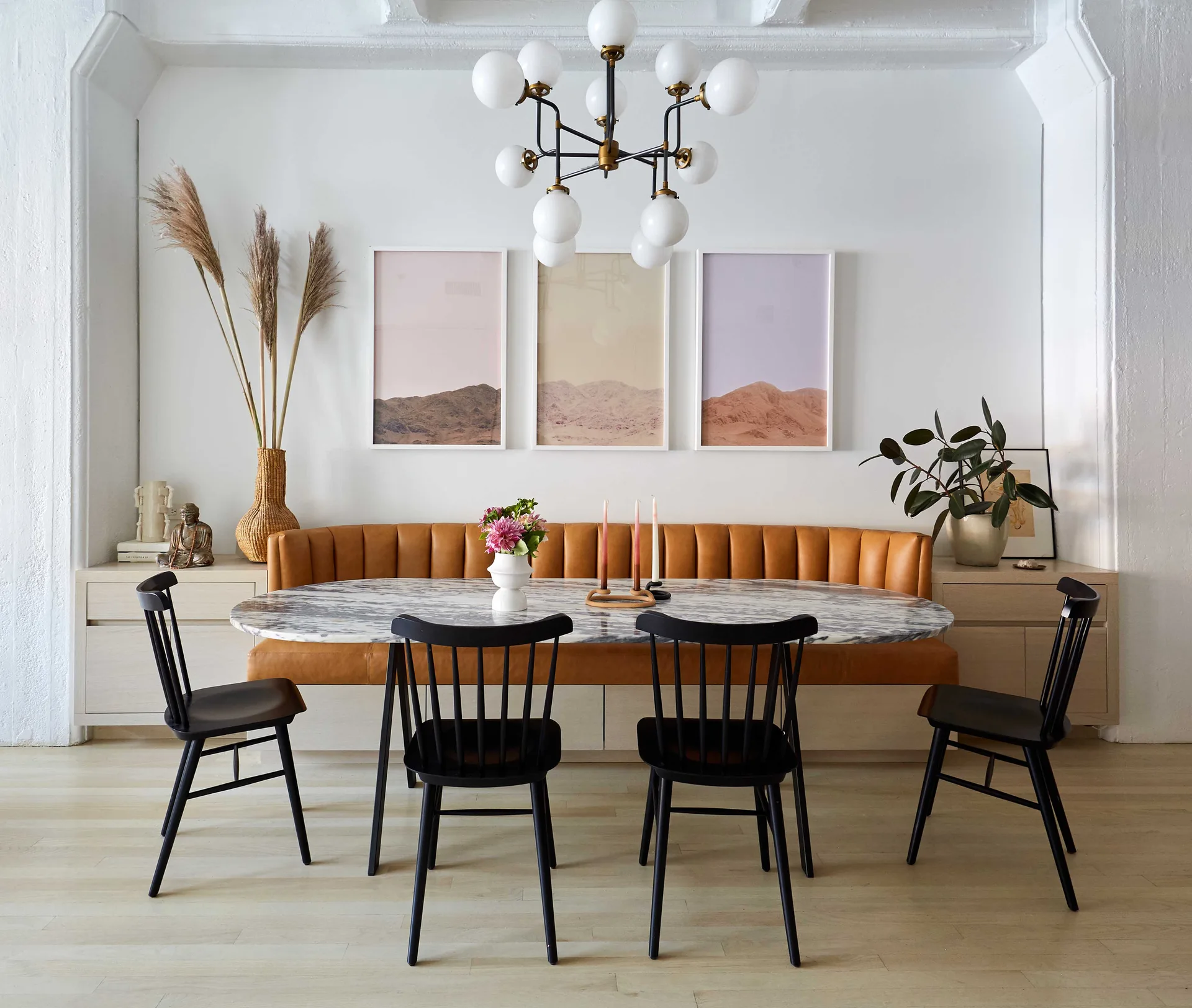 An oval grey-veined marble table is surrounded by black spindle chairs and a built-in tan leather banquette beneath a modern globe chandelier.