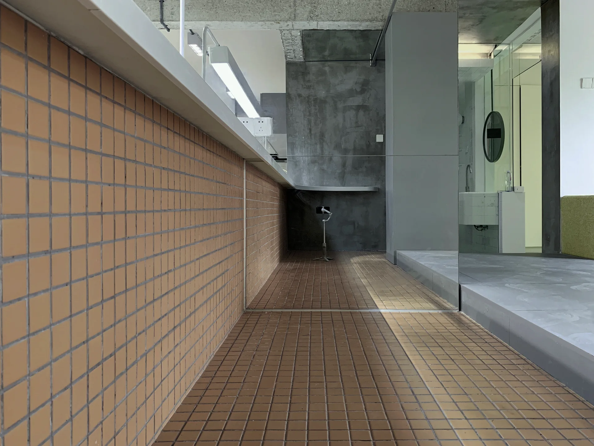 An industrial interior features a long counter and floor covered in orange square tiles, adjacent to a raw concrete wall and a large mirror.