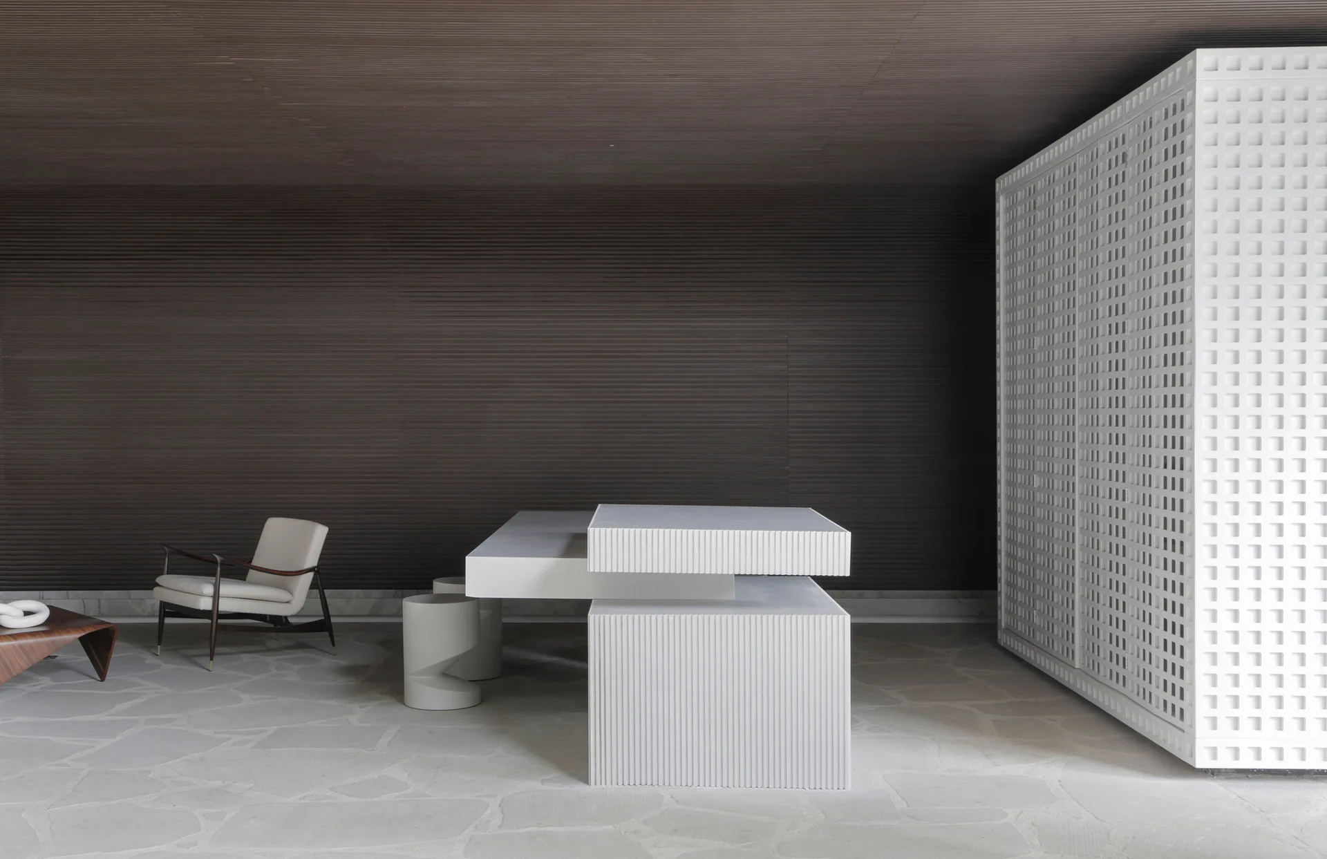 A minimalist lounge features dark slatted wood walls, a tiered white sculptural table, a white grid cabinet, and grey flagstone flooring.