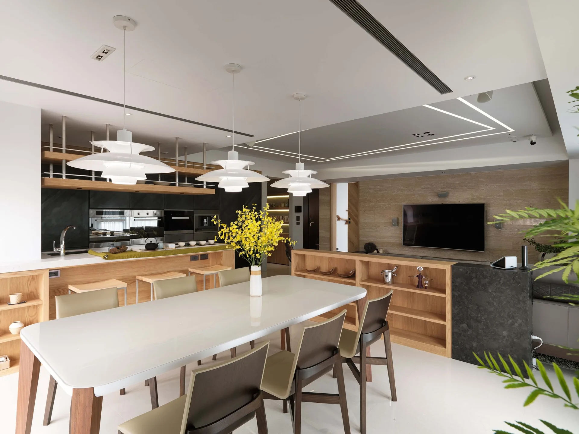 A modern dining area features a white table surrounded by chairs under three tiered pendant lights, with a low wooden console dividing the open-plan room.