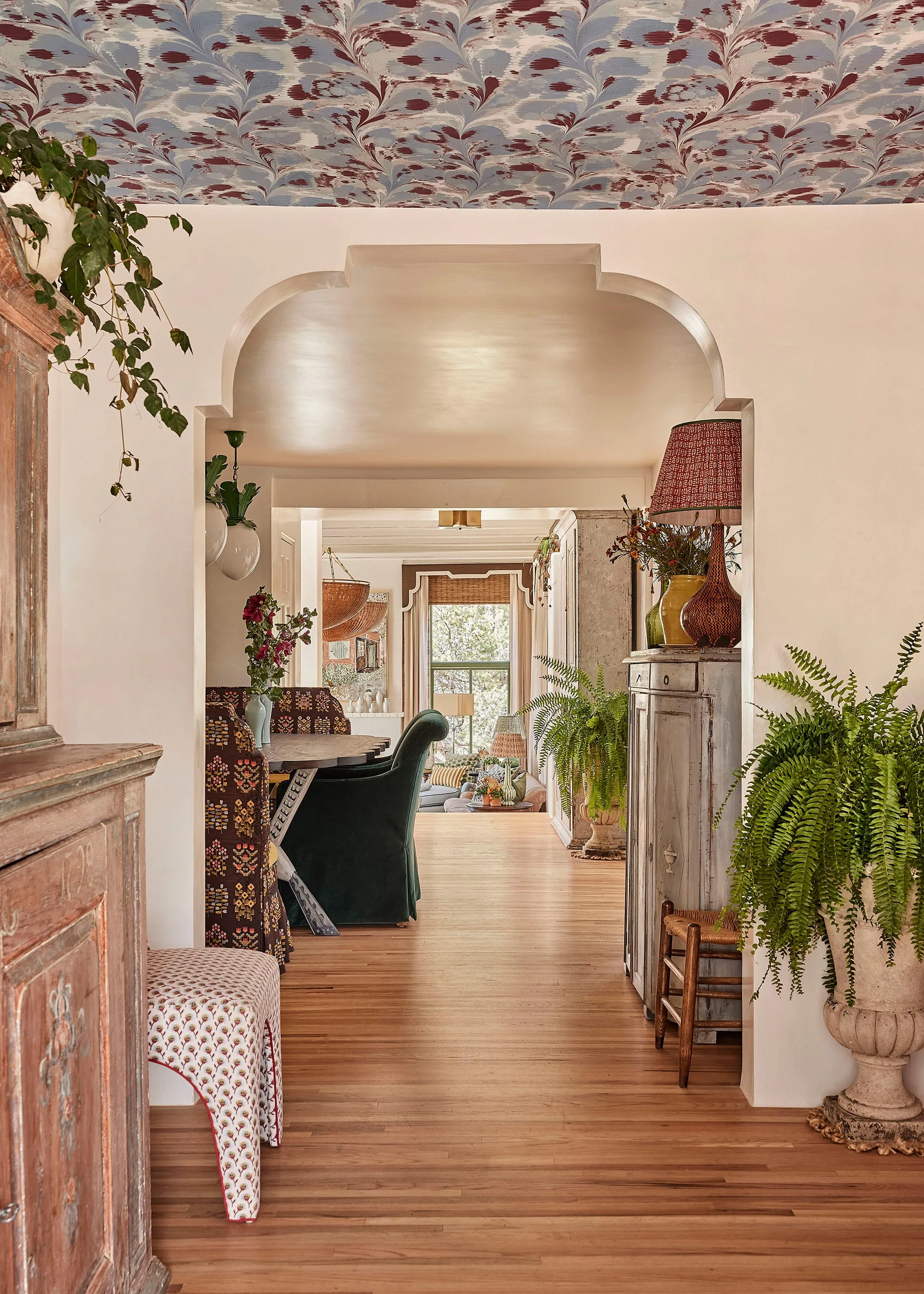An arched passage connects a hallway with marbled ceiling wallpaper and potted ferns to a dining area and living room.
