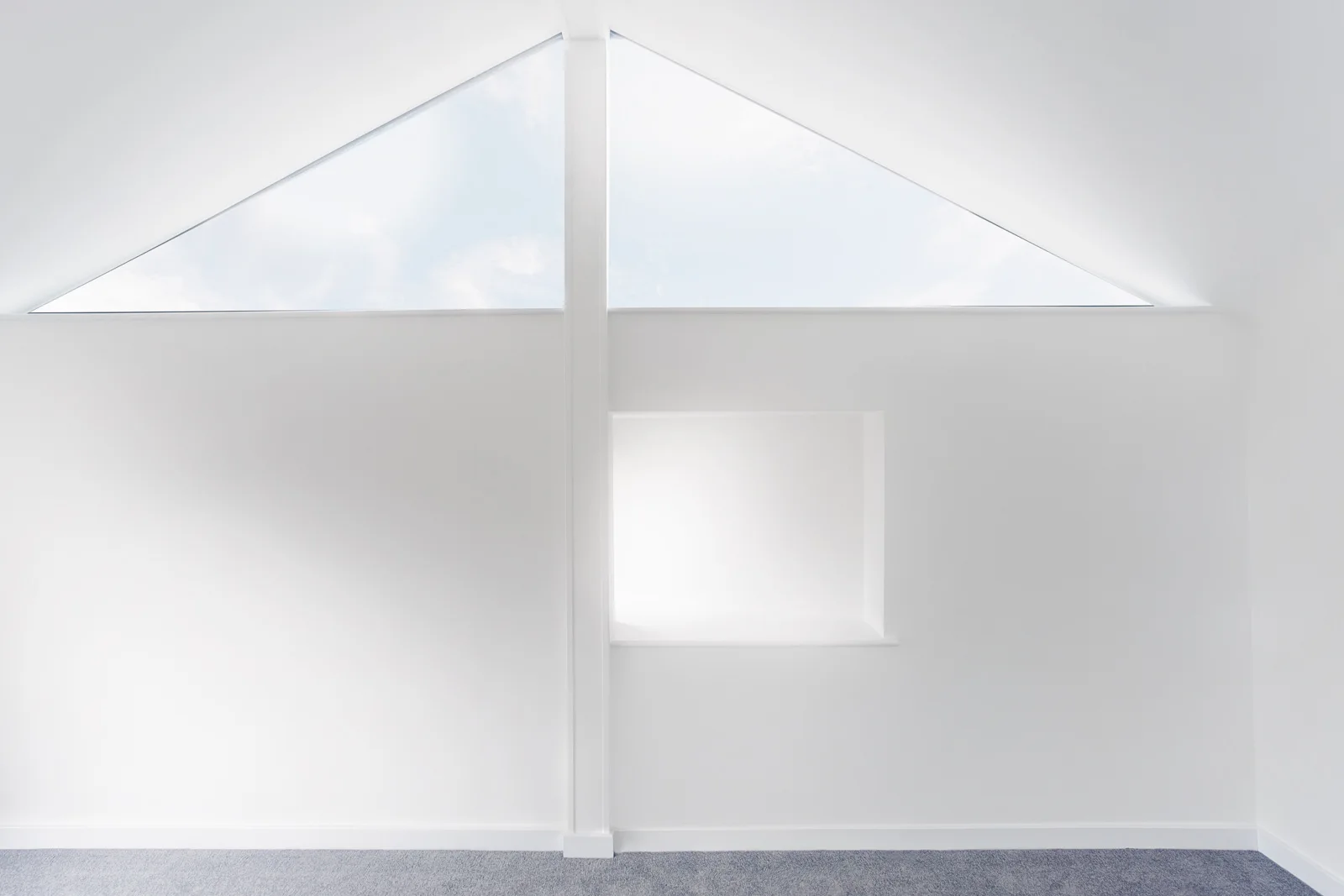 A minimalist white wall features a triangular gabled window near the sloped ceiling and a square recessed alcove above a grey carpeted floor.