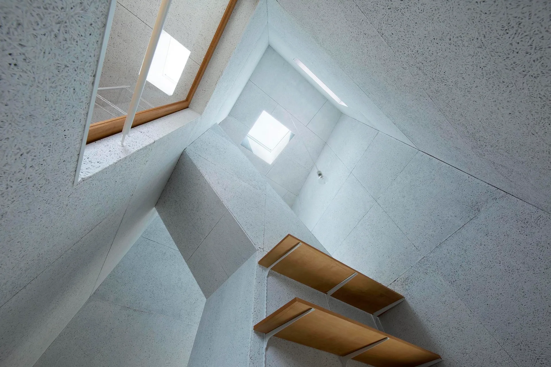 A low-angle view of floating wooden stair treads mounted on textured grey paneled walls under square skylights.