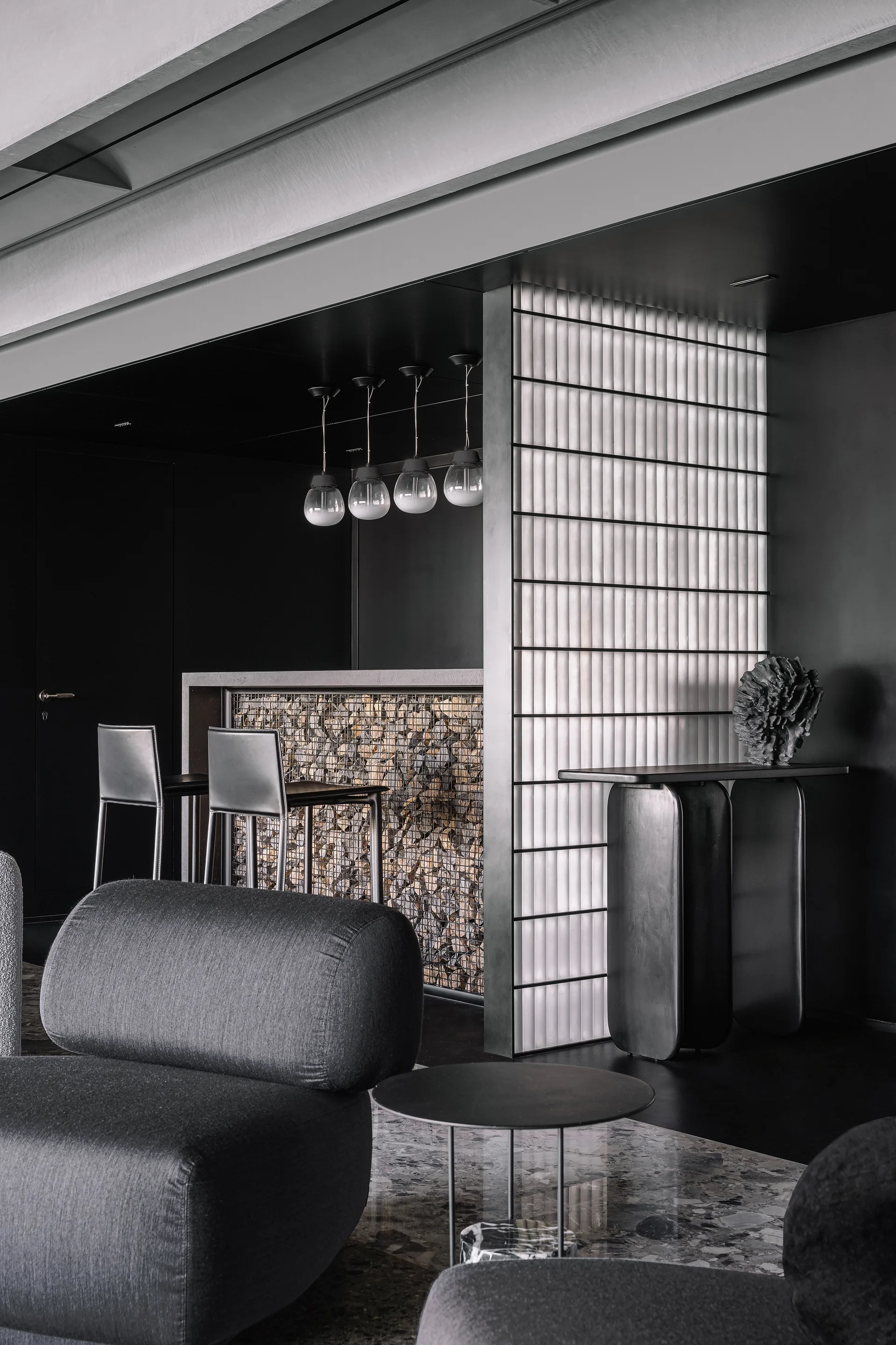 A dark moody lounge featuring grey upholstered chairs, a backlit grid wall partition, and a bar counter with a stone-filled metal cage base.
