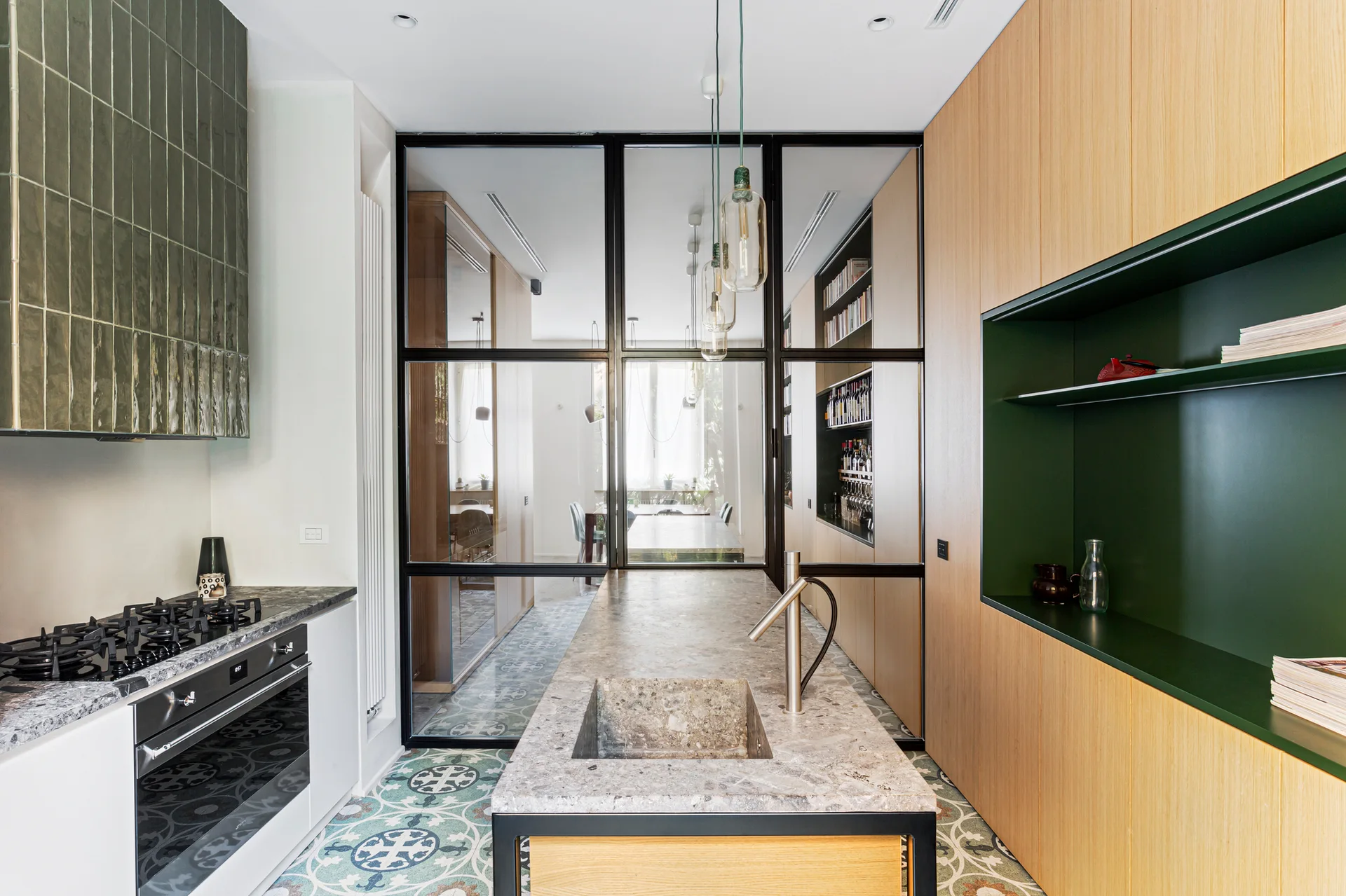 A modern kitchen with a stone-topped island, light wood cabinetry, a glass partition wall, and patterned floor tiles.