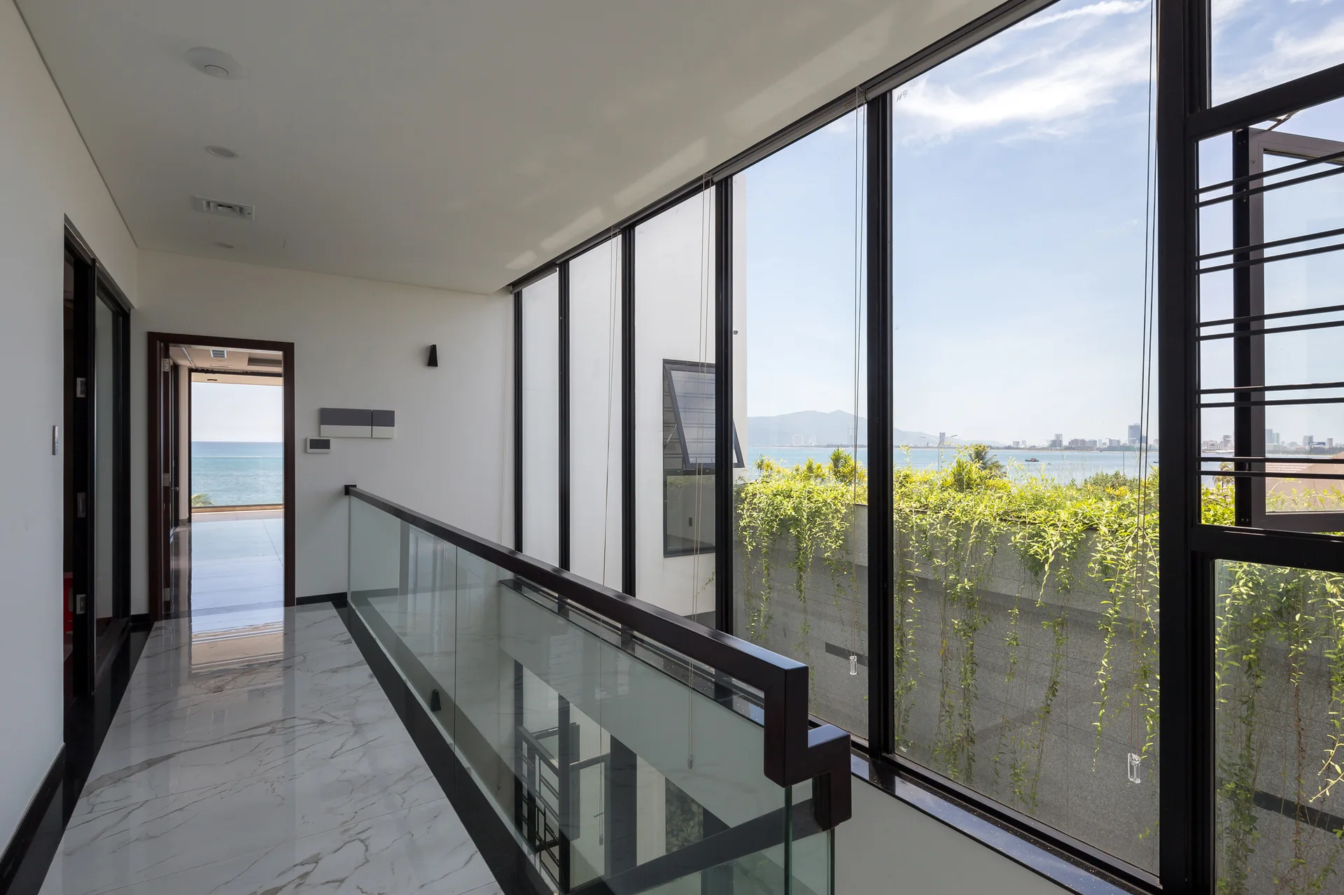 A modern hallway features polished white marble flooring, a glass railing, and large black-framed windows overlooking an ocean view.