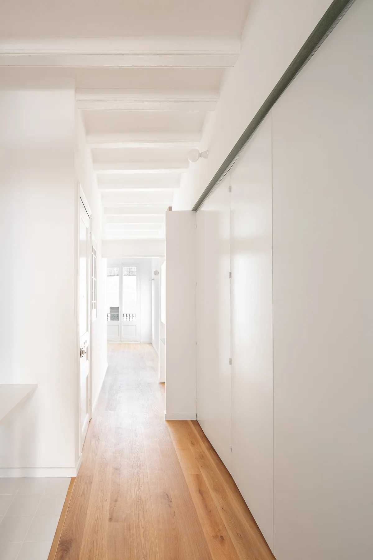 A bright minimalist hallway featuring light wood flooring, white-painted exposed ceiling beams, and flush white wall cabinets.
