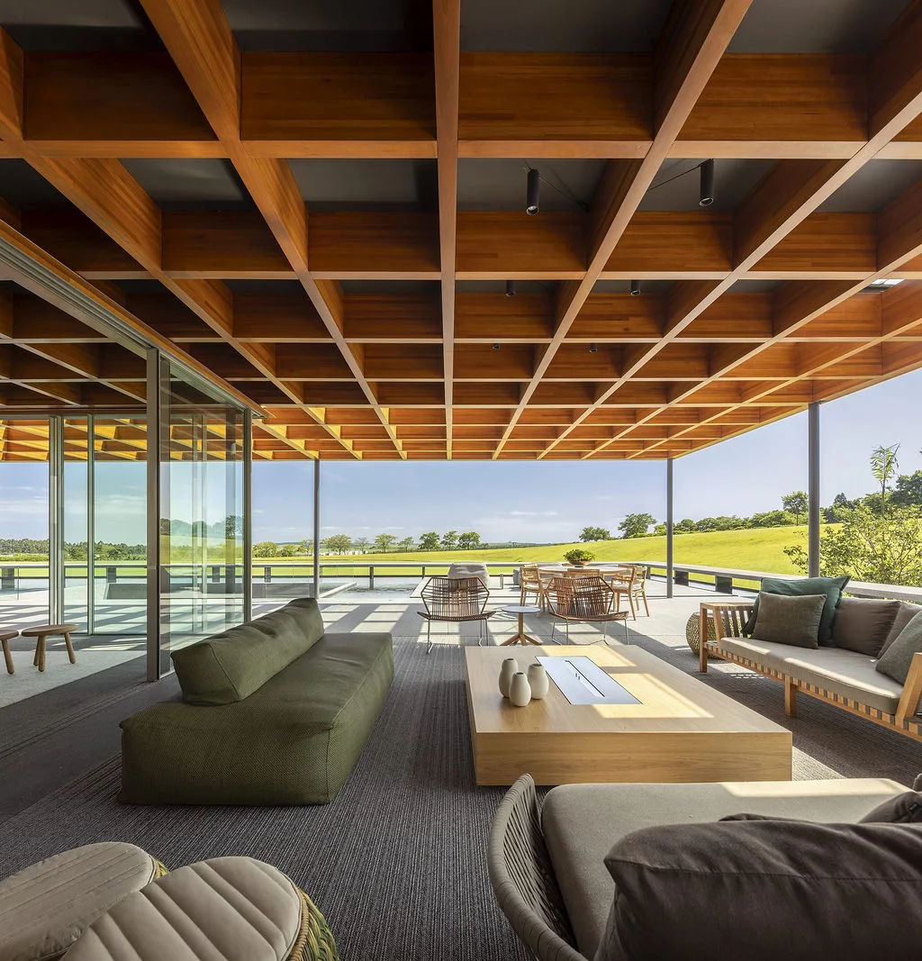 A spacious covered patio features a massive wooden grid coffered ceiling over olive green sofas and a low wooden coffee table with a grassy field view.